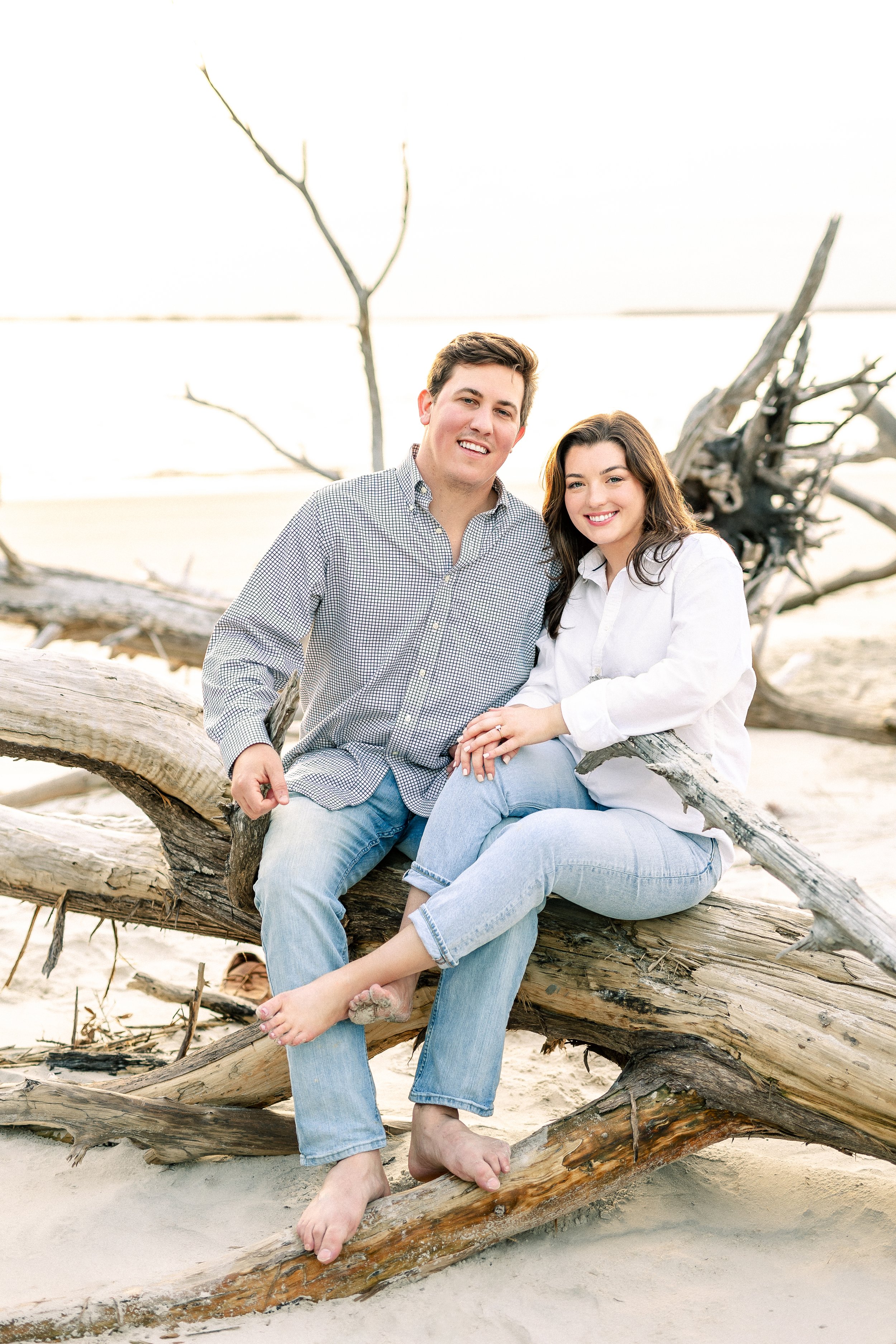 A young man and woman sitting on a large piece of driftwood on a sandy beach, smiling at the camera. The background features a driftwood tree and overcast sky.
