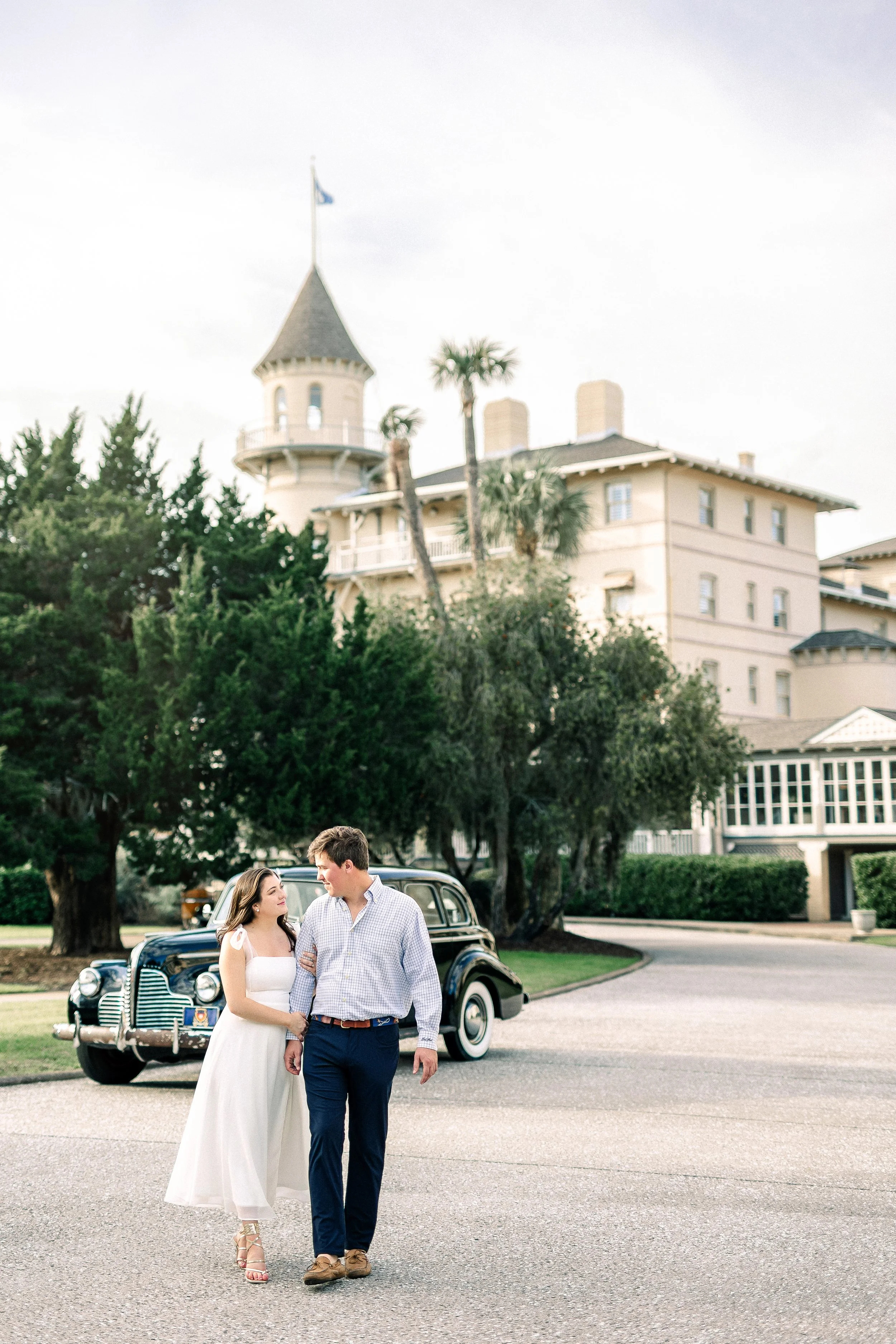 A couple is walking and talking in front of a vintage black car, with a large mansion and palm trees in the background.