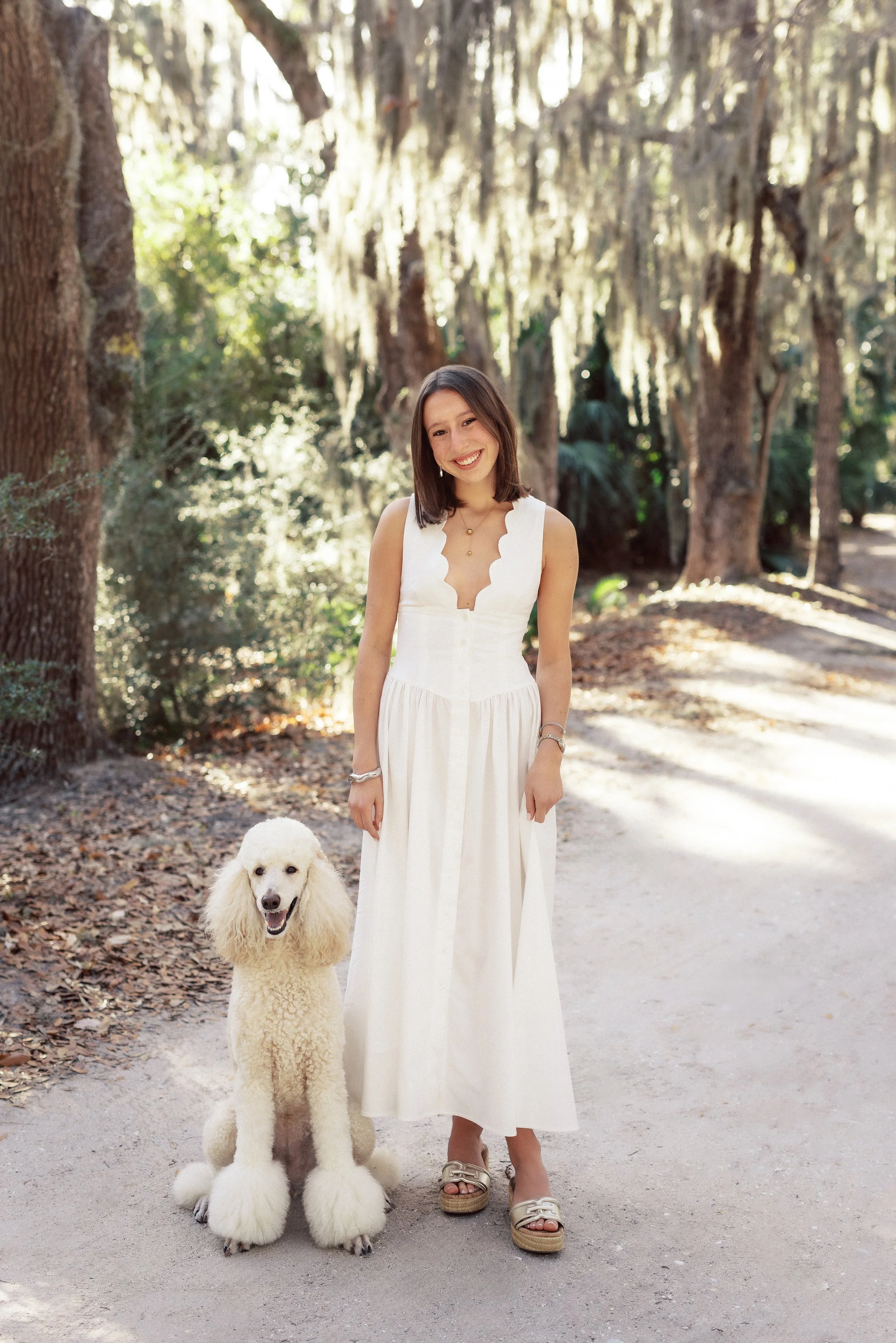 A woman in a white dress standing outdoors on a dirt path with a white poodle dogs. The background has tall trees and sunlight filtering through the leaves.