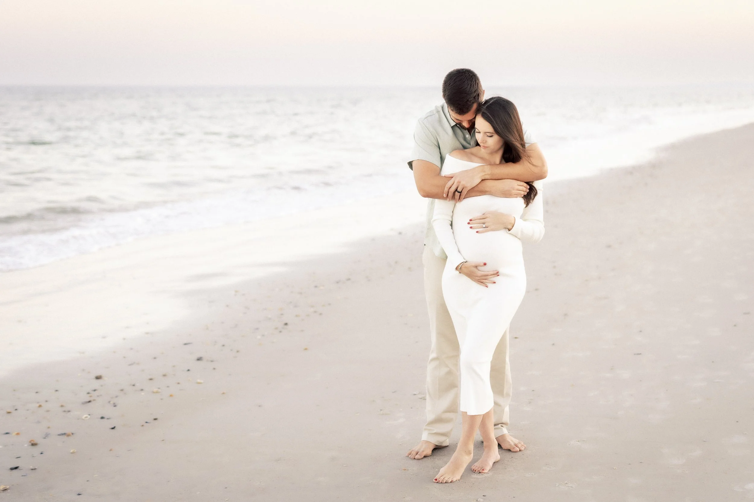 A couple, dressed in light-colored clothing, embracing on a sandy beach near the water with gentle waves.