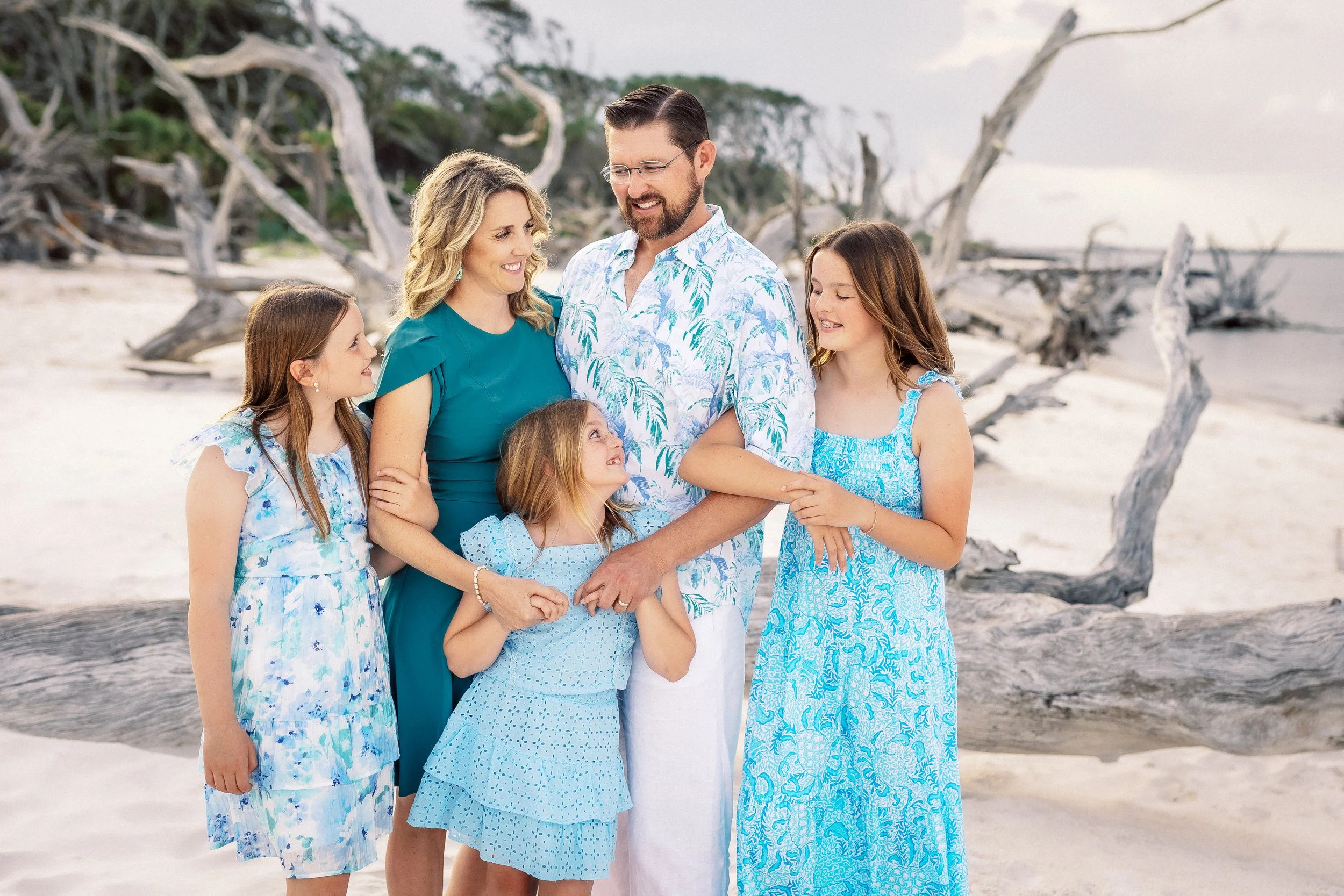 A family of six standing on a beach, smiling, with driftwood and a cloudy sky in the background.