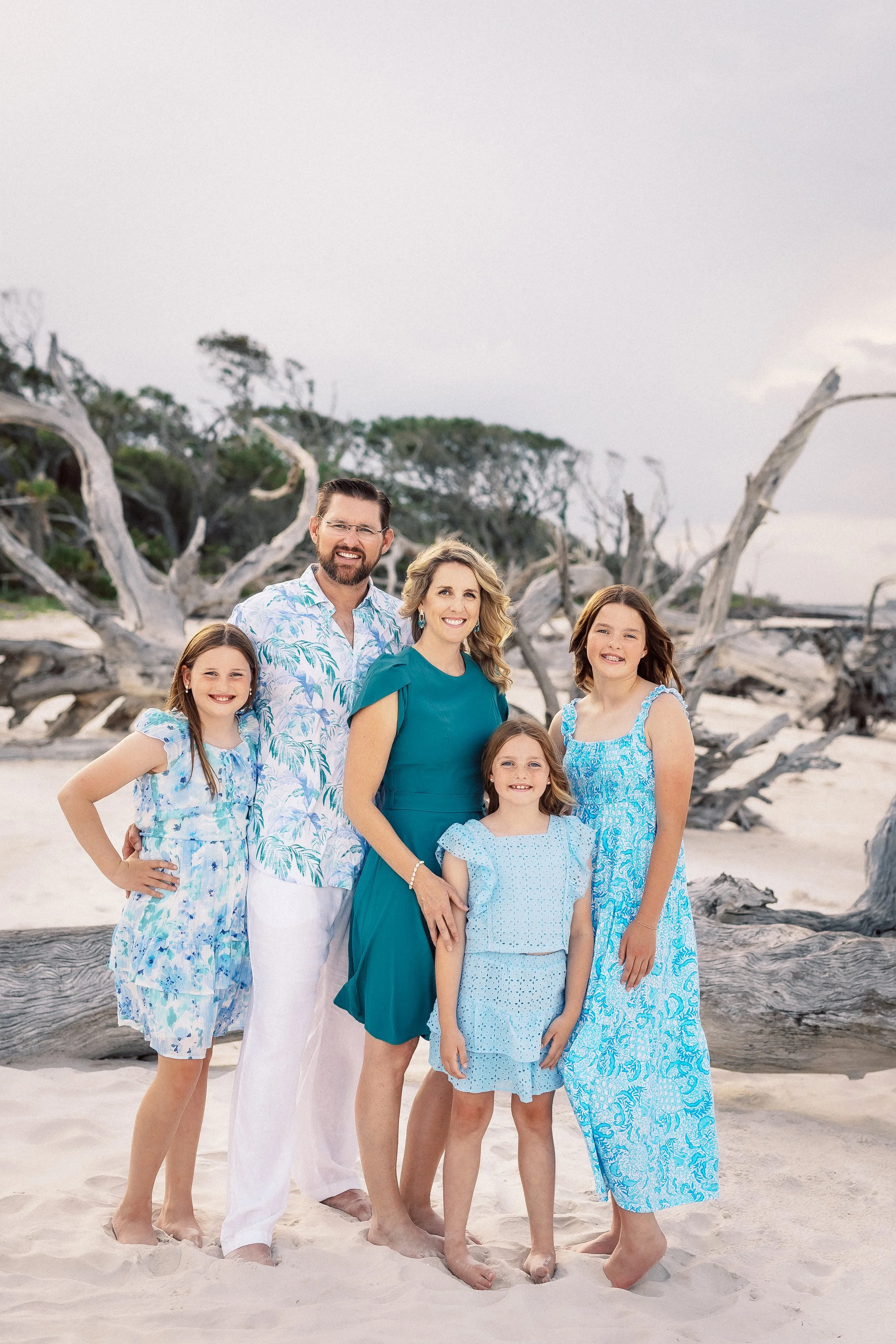 A family of seven standing on a sandy beach with driftwood and trees in the background, smiling for the photo. They are wearing summer clothing, mostly in shades of blue and white.
