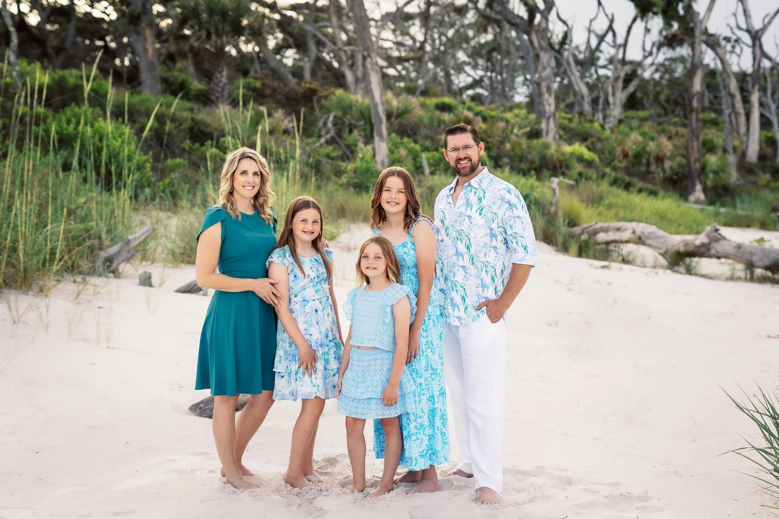 Family of six standing on a sandy beach with green vegetation and trees in the background. The family includes two adults and four children, all smiling and dressed in blue and white summer clothing.