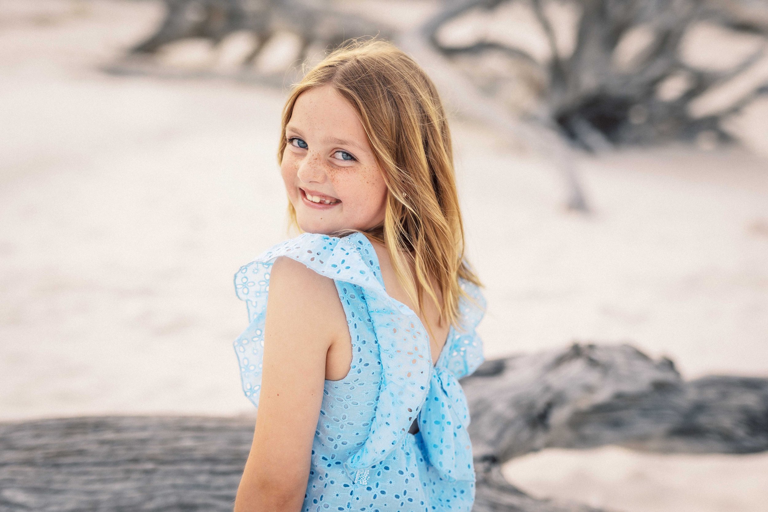 A young girl with red hair and freckles smiling and looking over her shoulder at the camera, wearing a blue dress with eyelet details, standing outdoors on a beach with driftwood and a tree in the background.