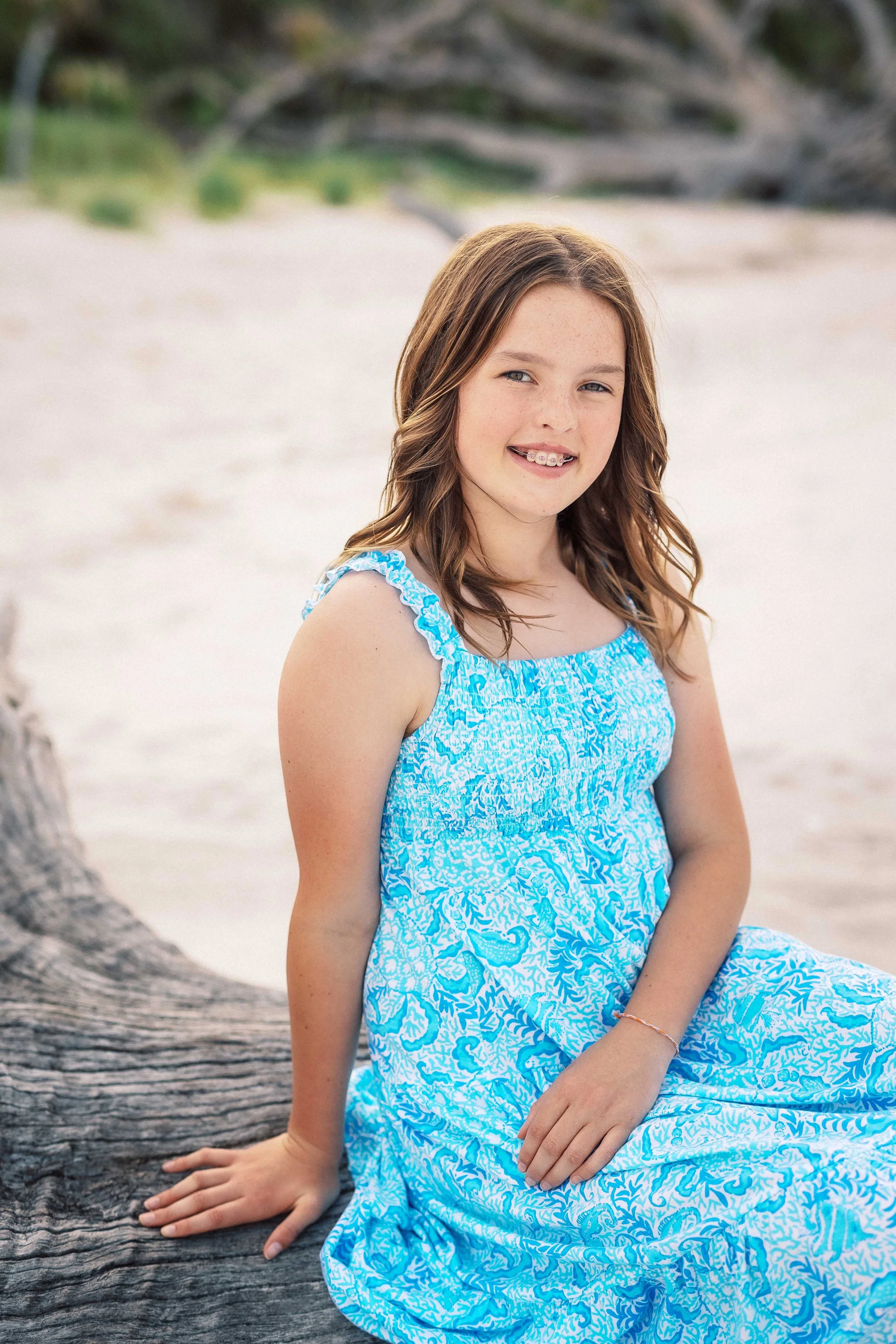 A young girl with brown hair wearing a blue and white patterned dress sitting on a large piece of driftwood on a beach.