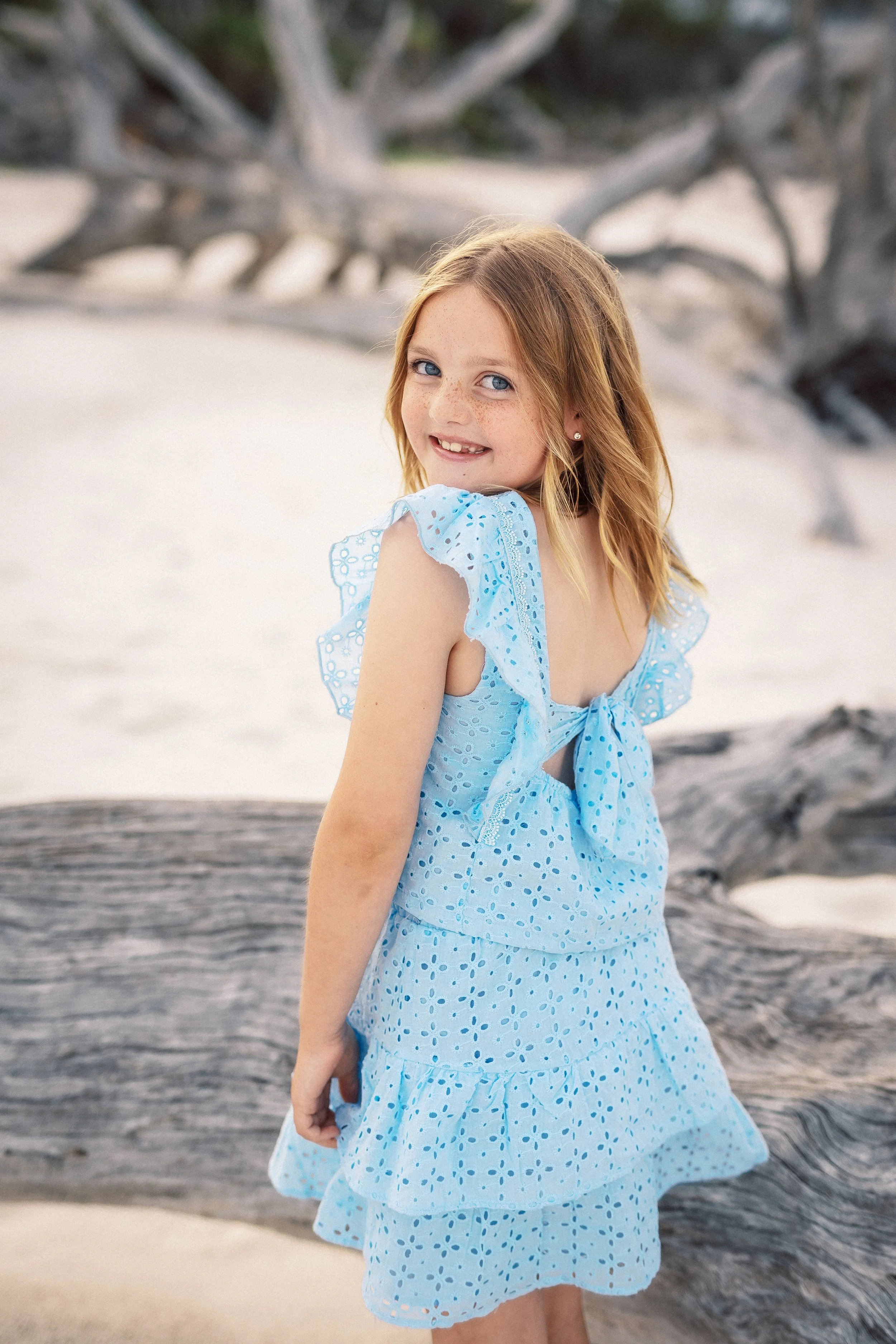 A young girl in a light blue dress with eyelet details standing outdoors near a piece of driftwood, with sandy ground and blurry trees in the background.