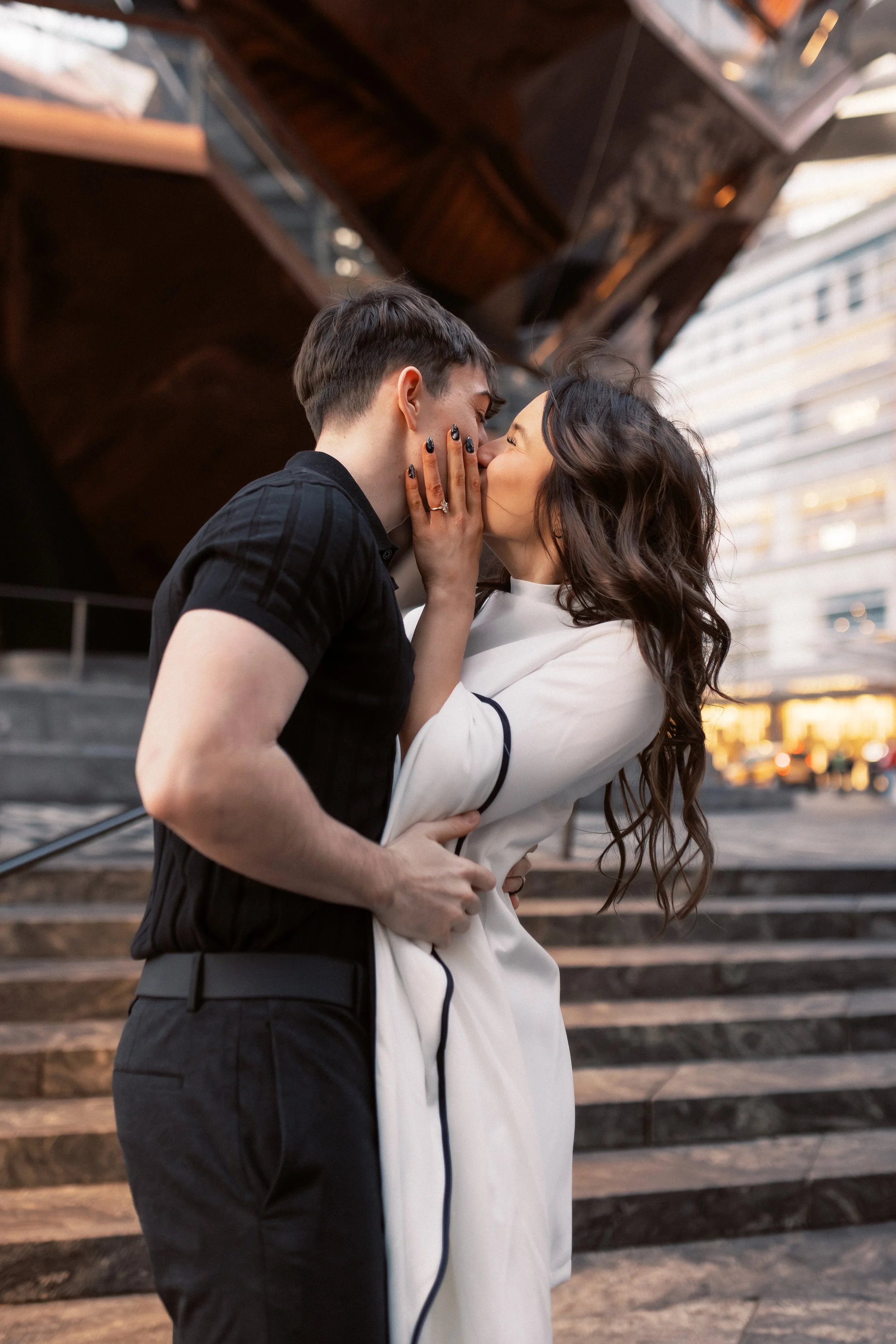 A couple is sharing a kiss on a city street at sunset, with a modern building in the background.