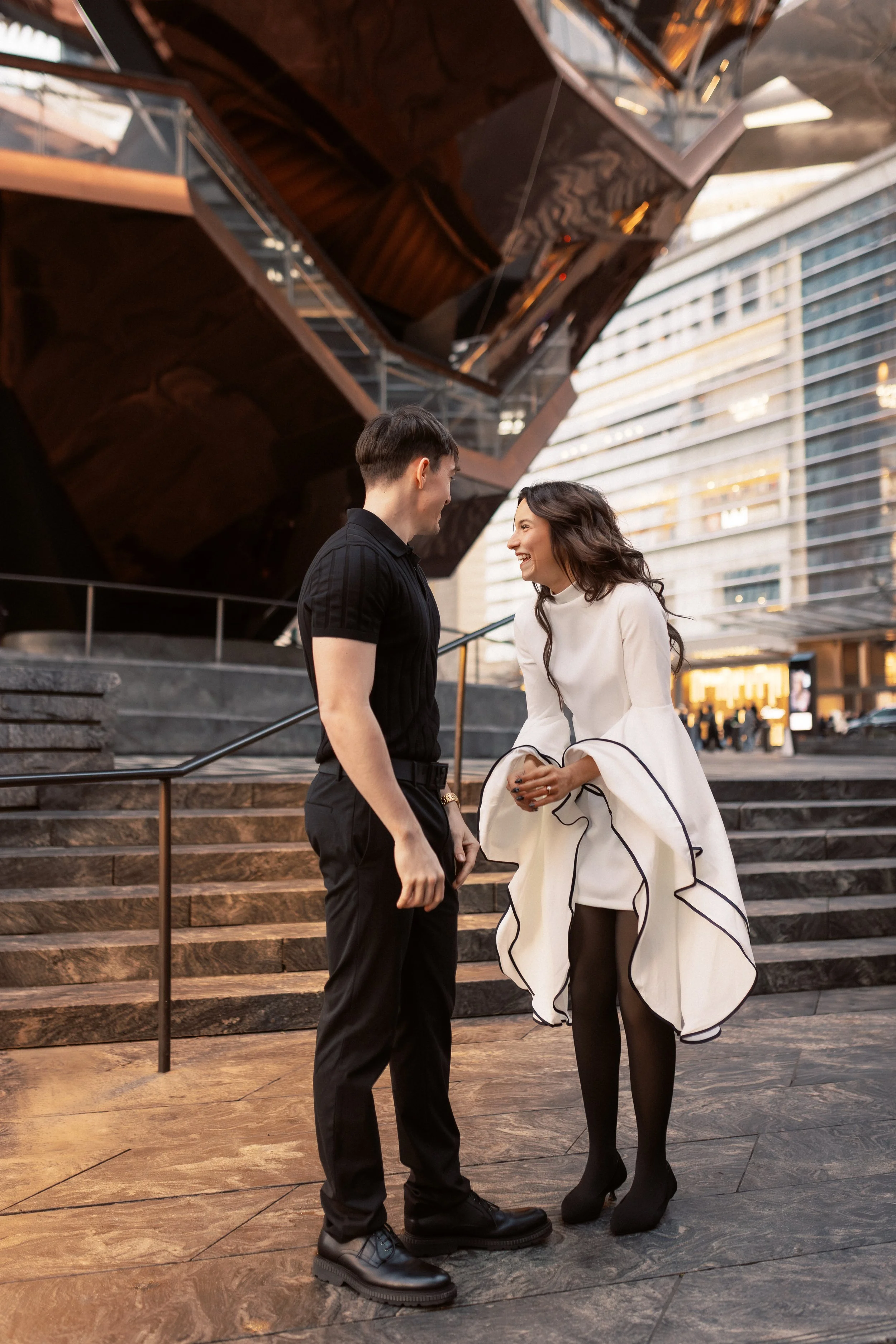 A young man and woman are standing on steps outside a modern building, smiling and facing each other. The woman is wearing a white dress with black trim and black tights, while the man is dressed in black with black shoes. The background features a g