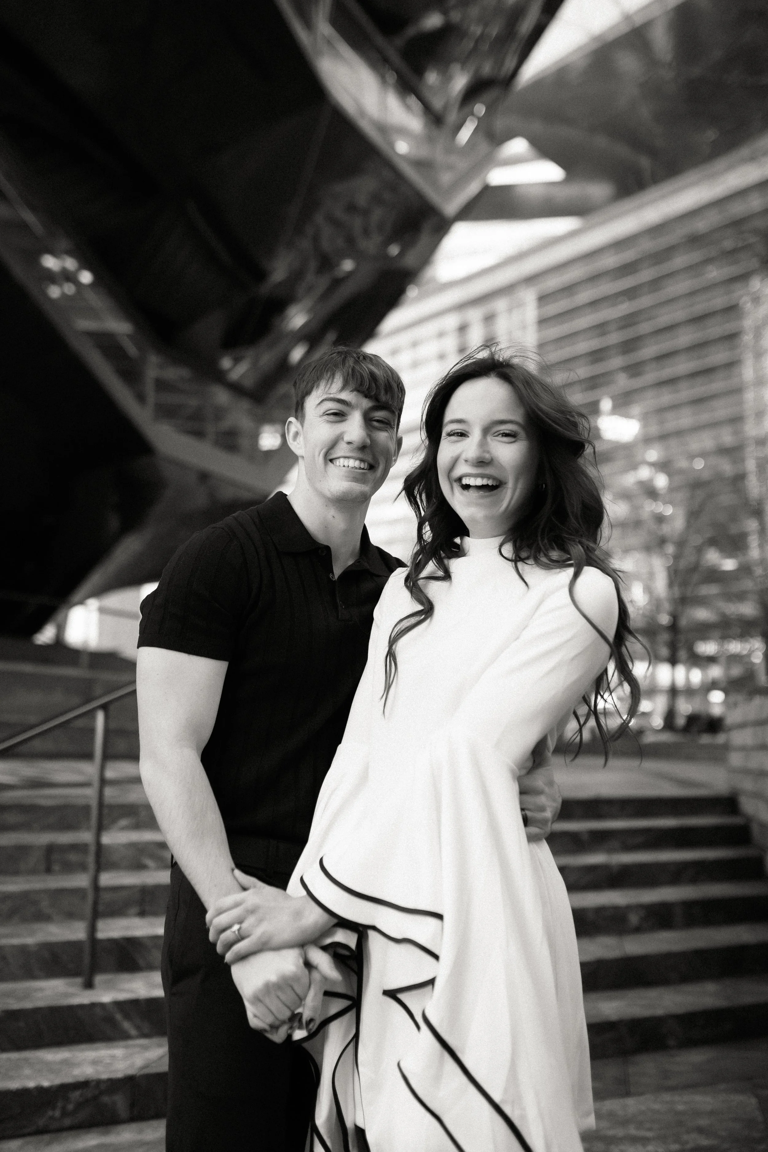 A young man and woman smiling and posing together outdoors in front of a modern building with stairs and metal railings. The photo is in black and white.