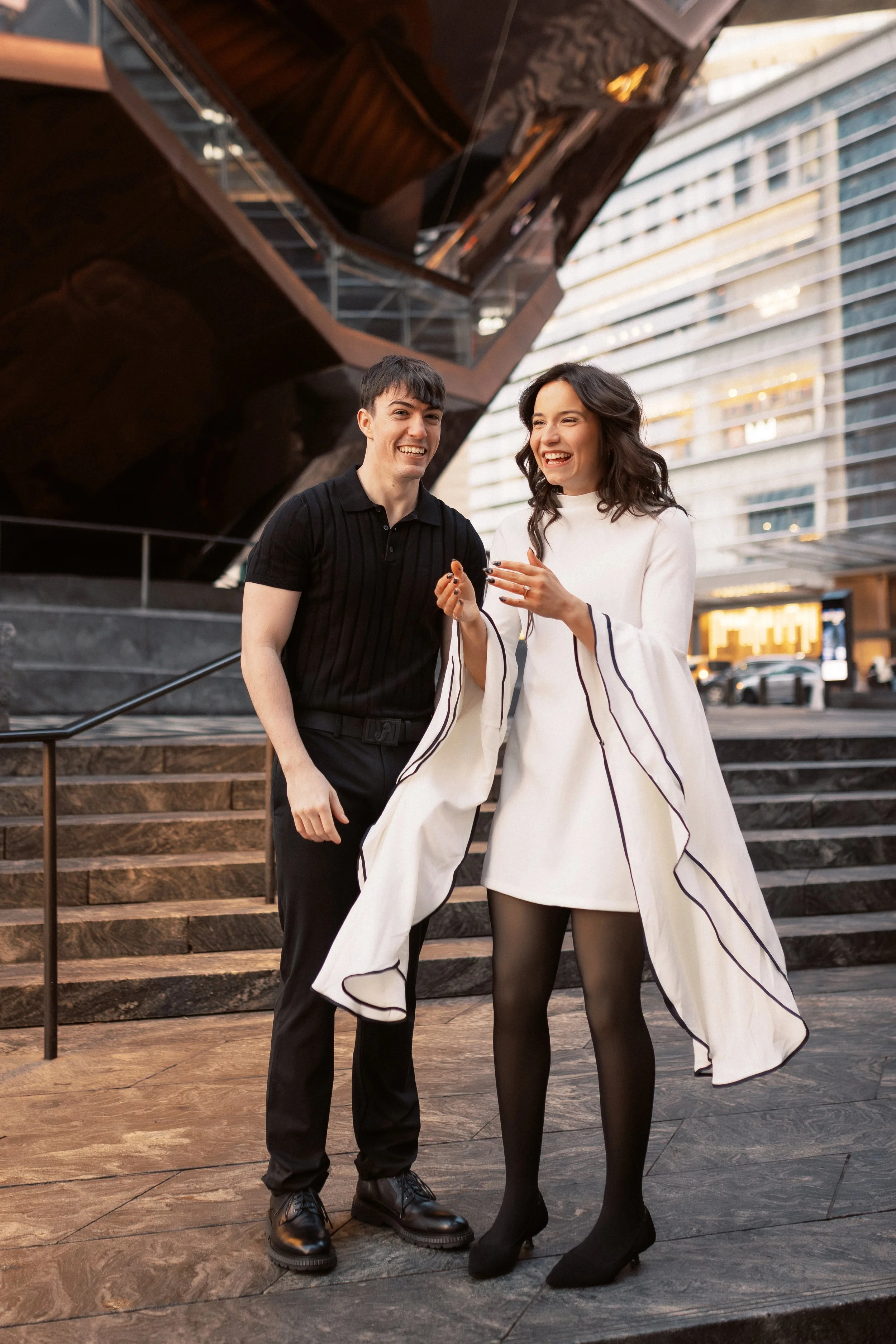 A man and a woman standing on stone steps outside a modern building, laughing and enjoying each other's company. The woman is dressed in a white dress with black stockings, and the man is wearing a black polo shirt and black pants.