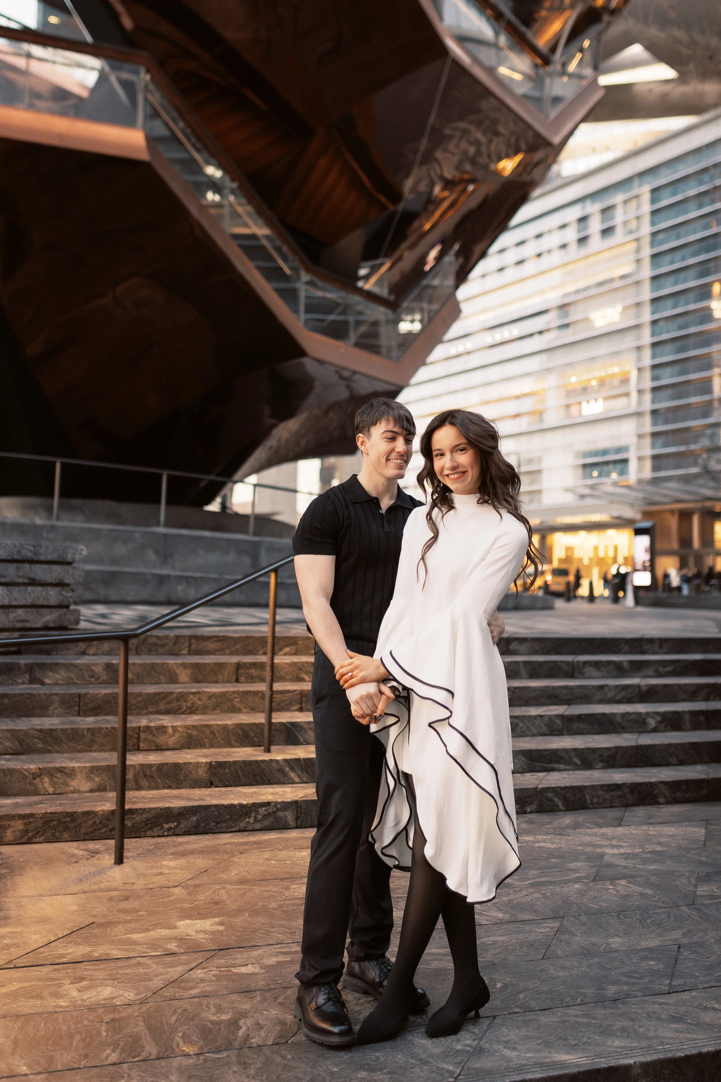 A smiling man and woman pose together outdoors in front of a modern building with large glass windows and metal panels. The man wears a black short-sleeve shirt and dark pants, and the woman wears a white dress with black trim and black tights, holdi
