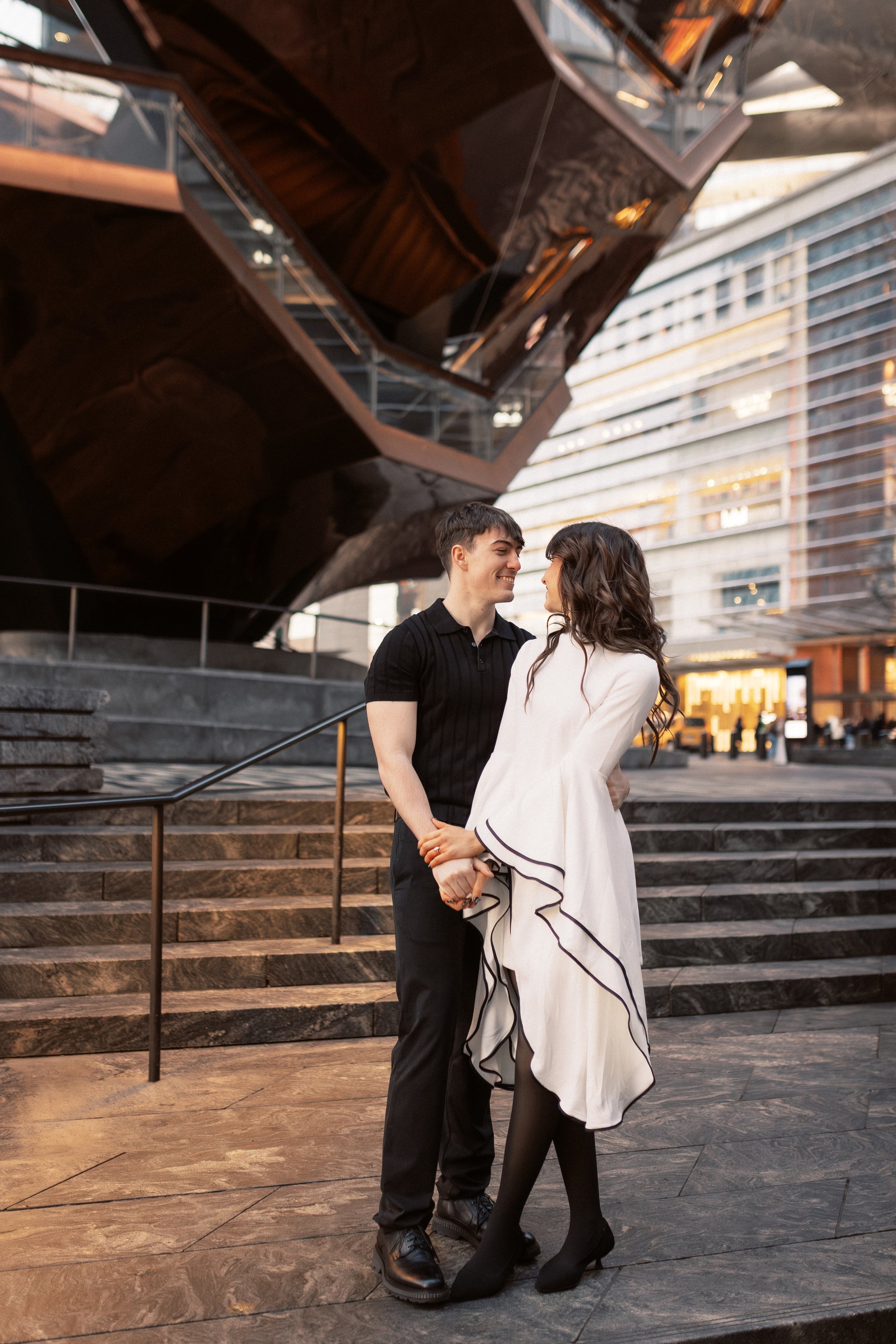 A young couple stands close together outdoors in front of a modern building with large glass windows and a staircase. They are smiling at each other, holding hands, with the man wearing a black polo shirt and black pants, and the woman wearing a whit