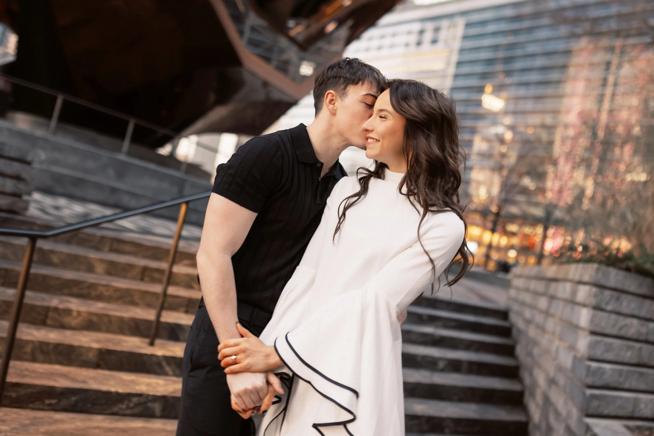 A young couple with light skin, standing outdoors in an urban setting, sharing a kiss and holding hands. The man has short dark hair and is wearing a black shirt, while the woman has long wavy dark hair and is dressed in a white top with black trim. 