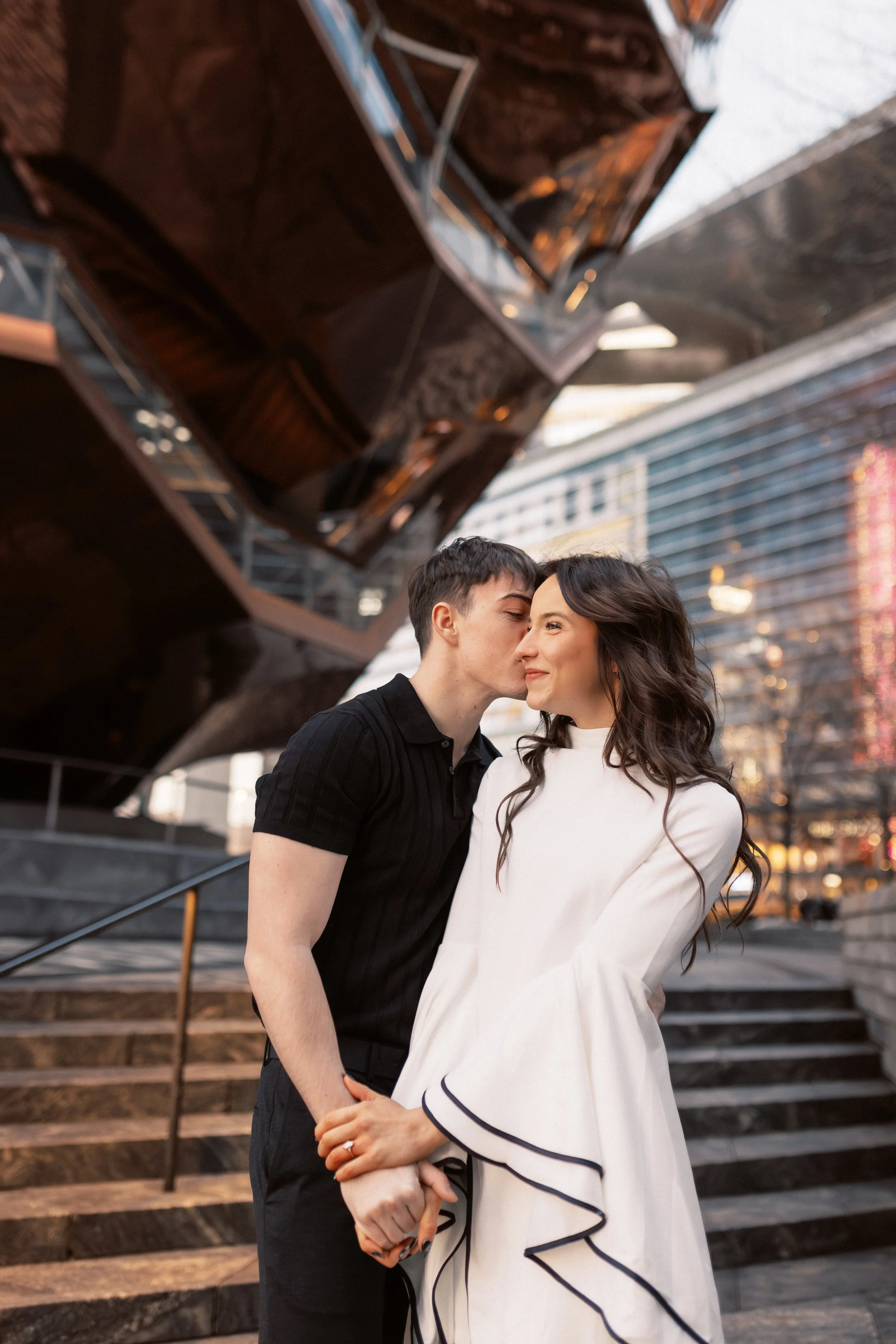 A young couple is standing close together in an urban setting with modern buildings in the background. The man is whispering or kissing the woman on her forehead, and they are holding hands. The woman has long dark hair and is wearing a white dress w