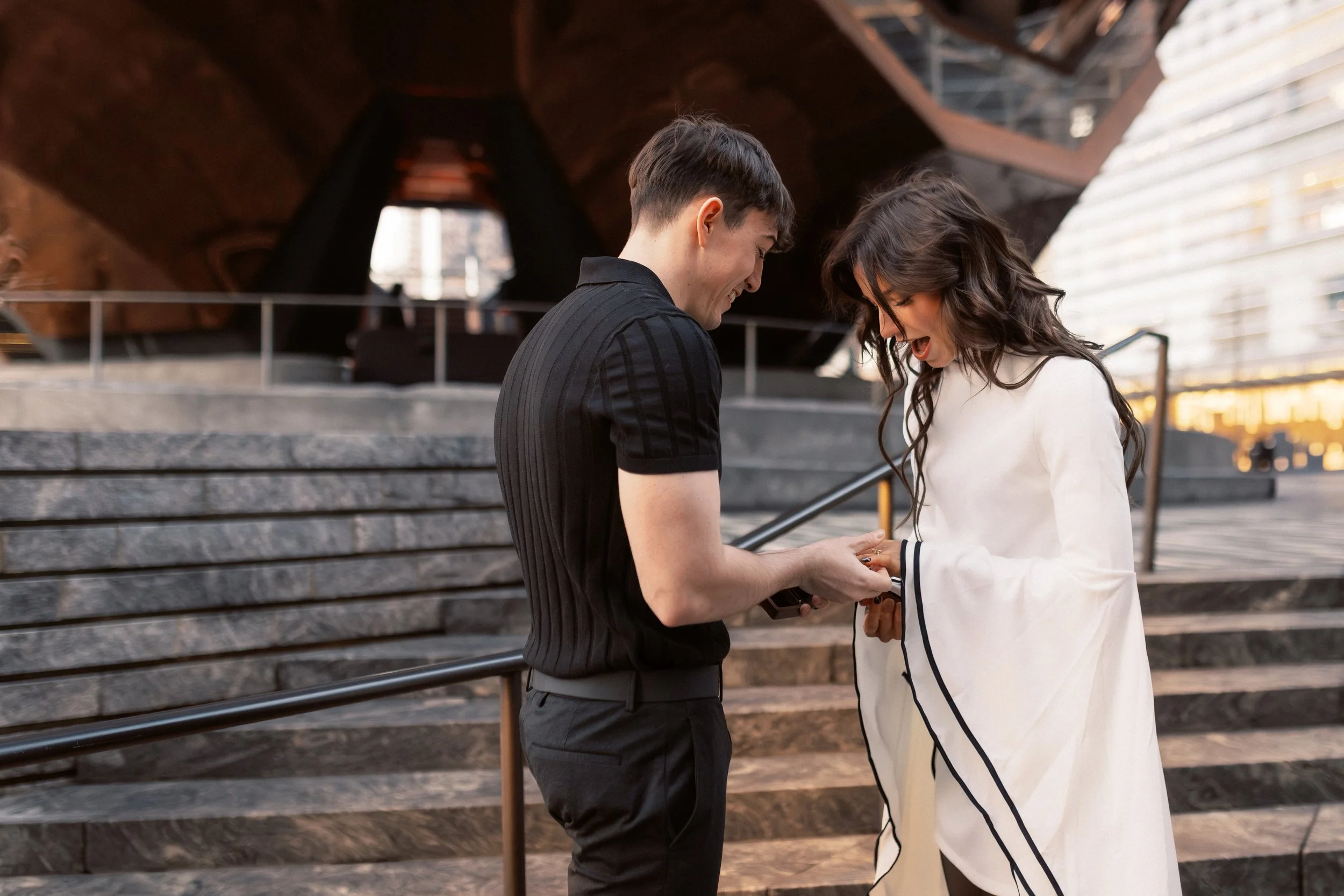A young man and woman sharing a joyful moment outdoors, with the man giving the woman a ring, in front of a set of stairs and a modern building.