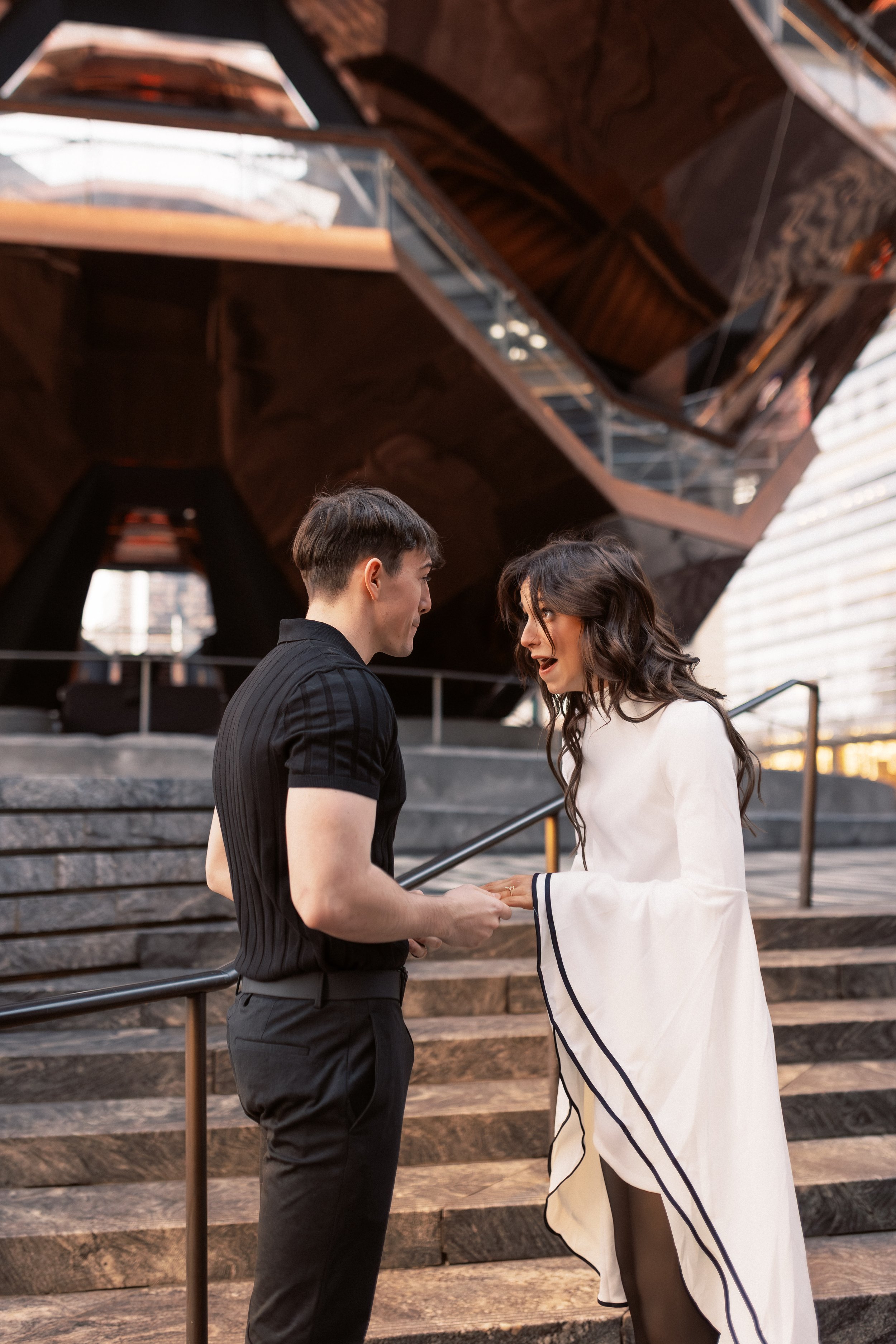 A man and woman are standing on steps outside at sunset, holding hands and looking into each other's eyes, with a modern building with a wood and metal structure behind them.