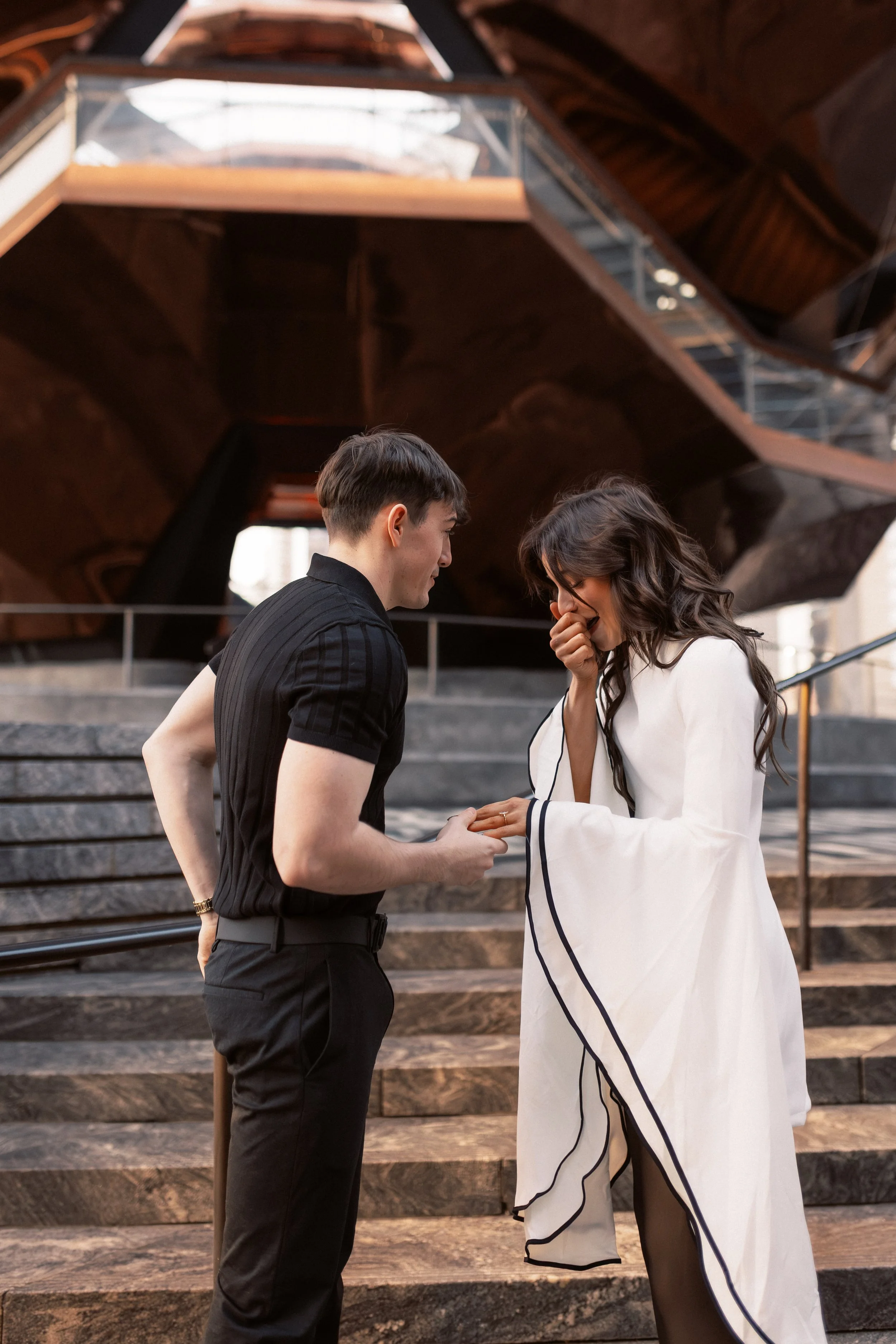 A man and a woman standing on steps outside, engaging in a conversation. The man is wearing a black striped shirt and black pants, while the woman is dressed in a white outfit with black trim, holding her hand near her face and smiling. Behind them i