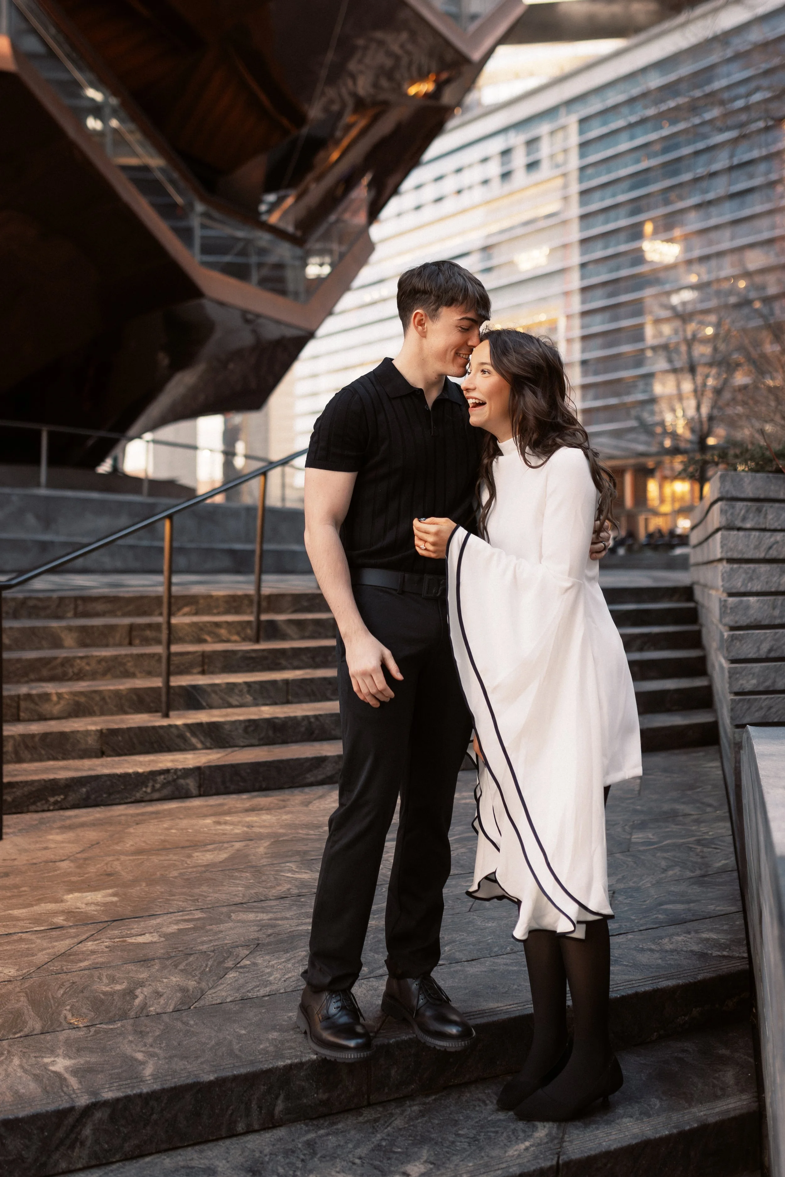 A young man and woman standing close together on outdoor stairs, smiling and sharing a happy moment, with modern office buildings in the background during evening.