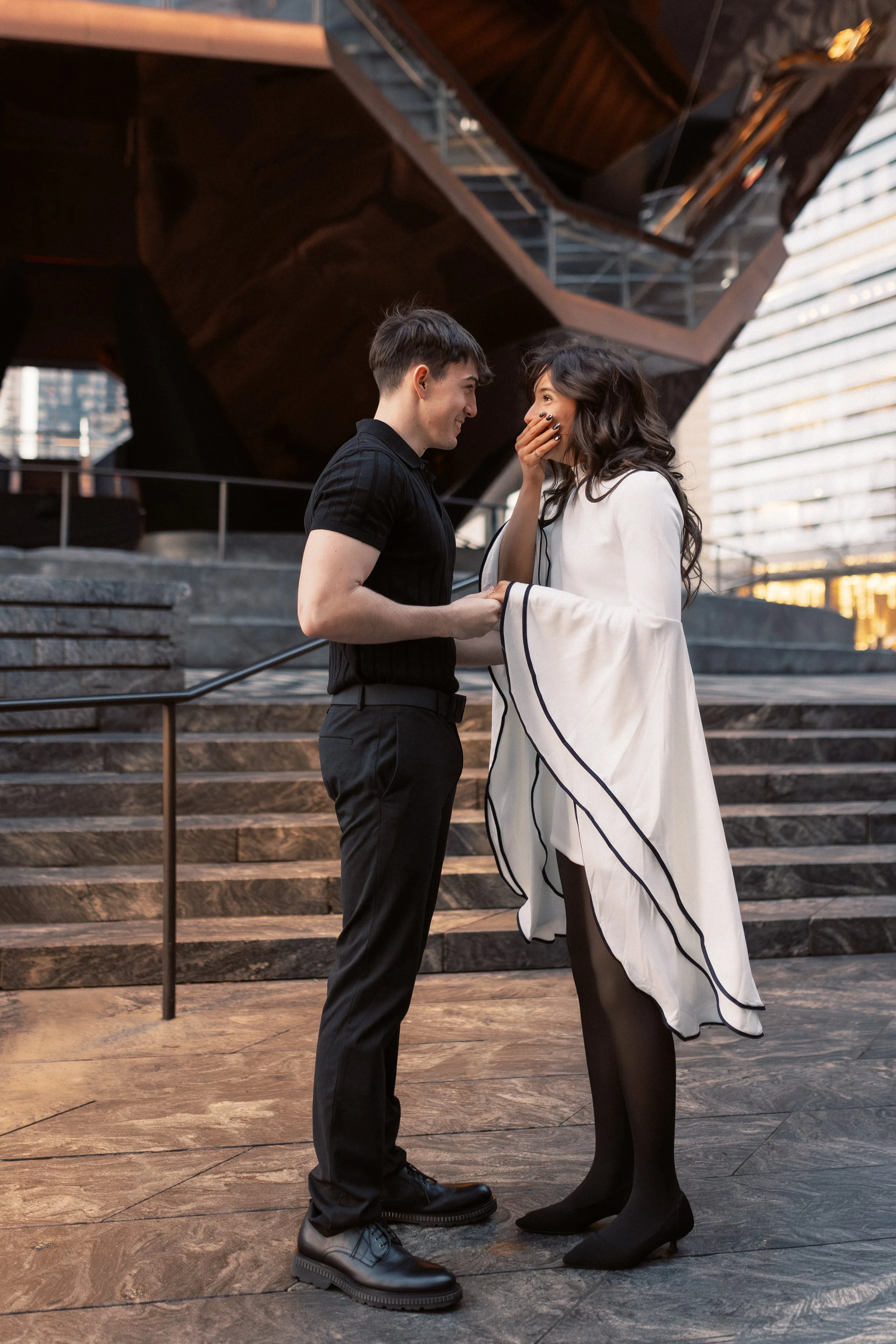 A man in black shirt and pants holding hands with a woman in a white coat with black stripes, who is covering her mouth with her hand, standing on a staircase outside an urban building with modern architecture and glass windows.