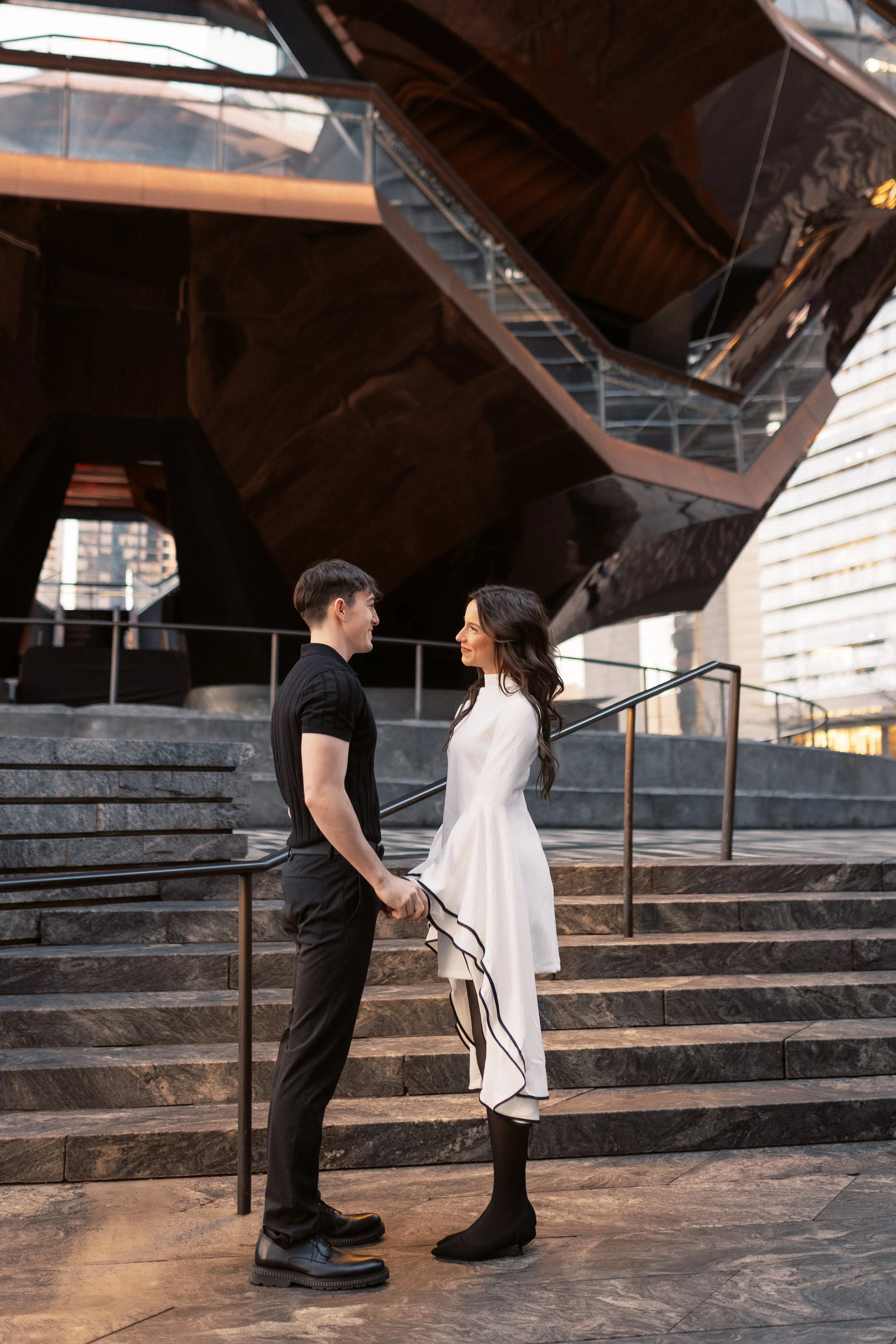 A couple is standing on the stairs in front of a modern architectural building, holding hands and looking into each other's eyes. The man is wearing a black outfit, and the woman is wearing a white dress with black accents. The building features dark
