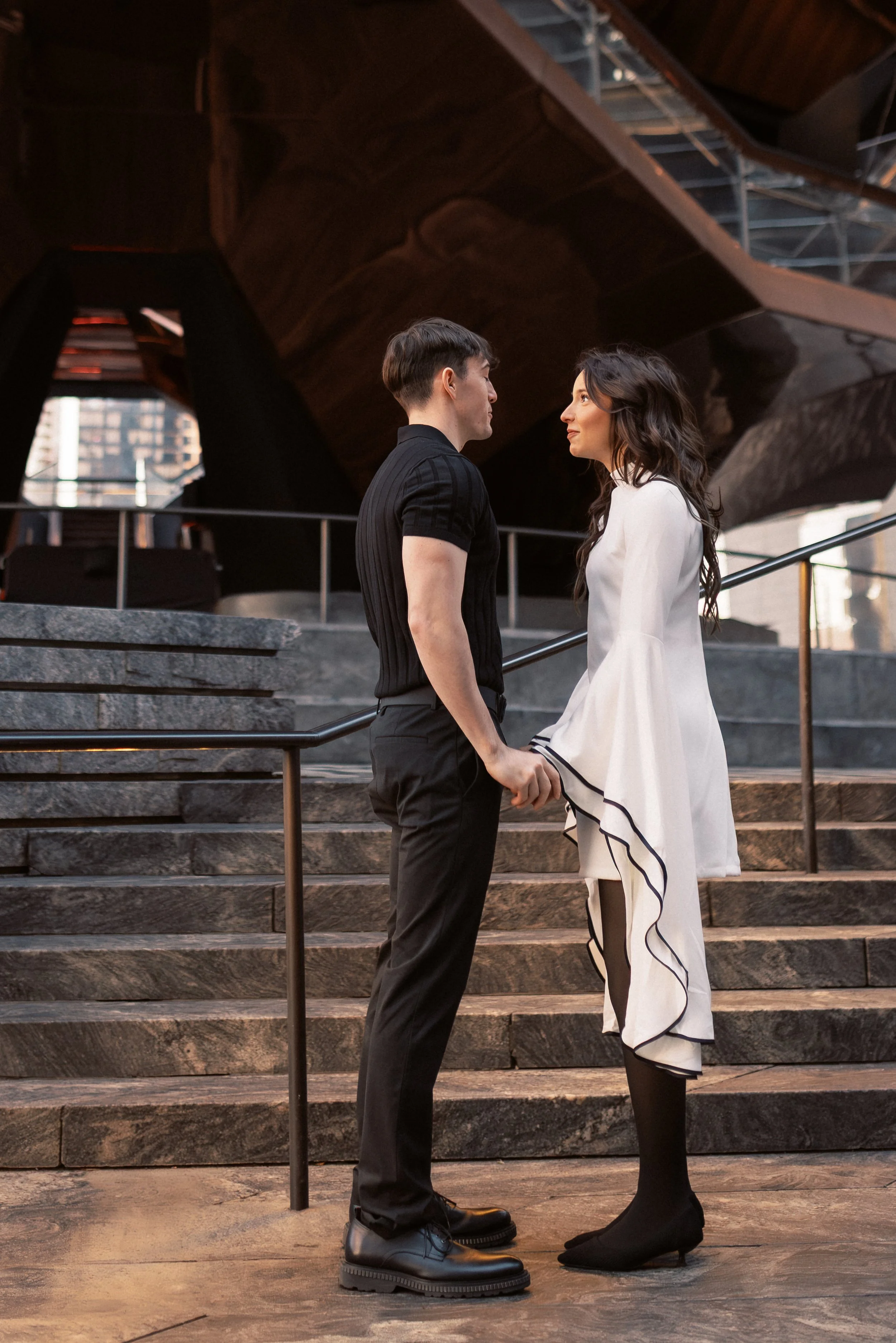 A young man and woman are standing on stairs, holding hands, facing each other and gazing into each other's eyes. The man is wearing a black short-sleeve shirt, black pants, and black boots. The woman is dressed in a white dress with black trim, blac