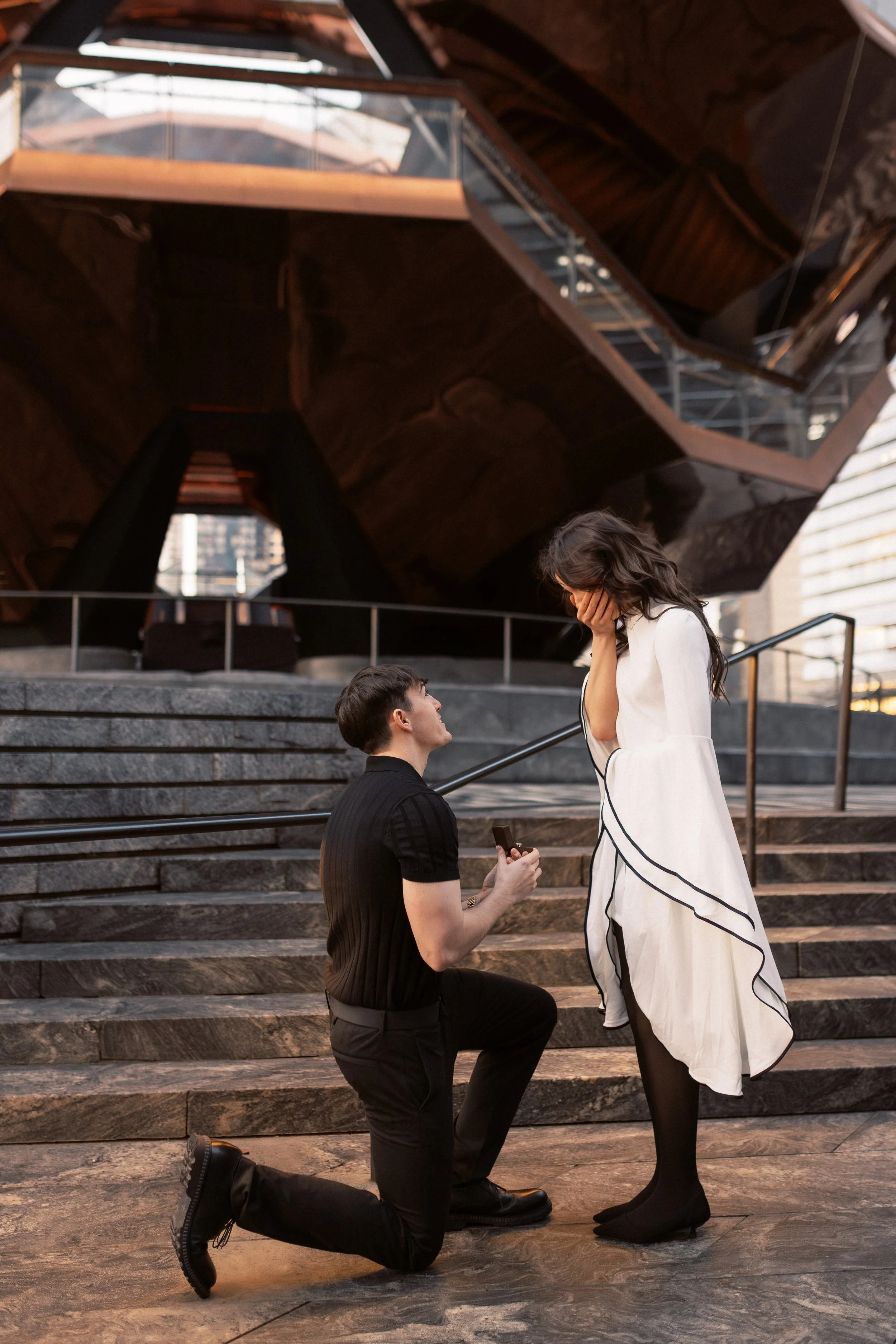 A man on one knee proposing marriage to a woman in front of a modern architectural building. The woman looks surprised and emotional, covering her face with one hand. The man is holding a small box, likely a ring. They are on stone steps, and the sce