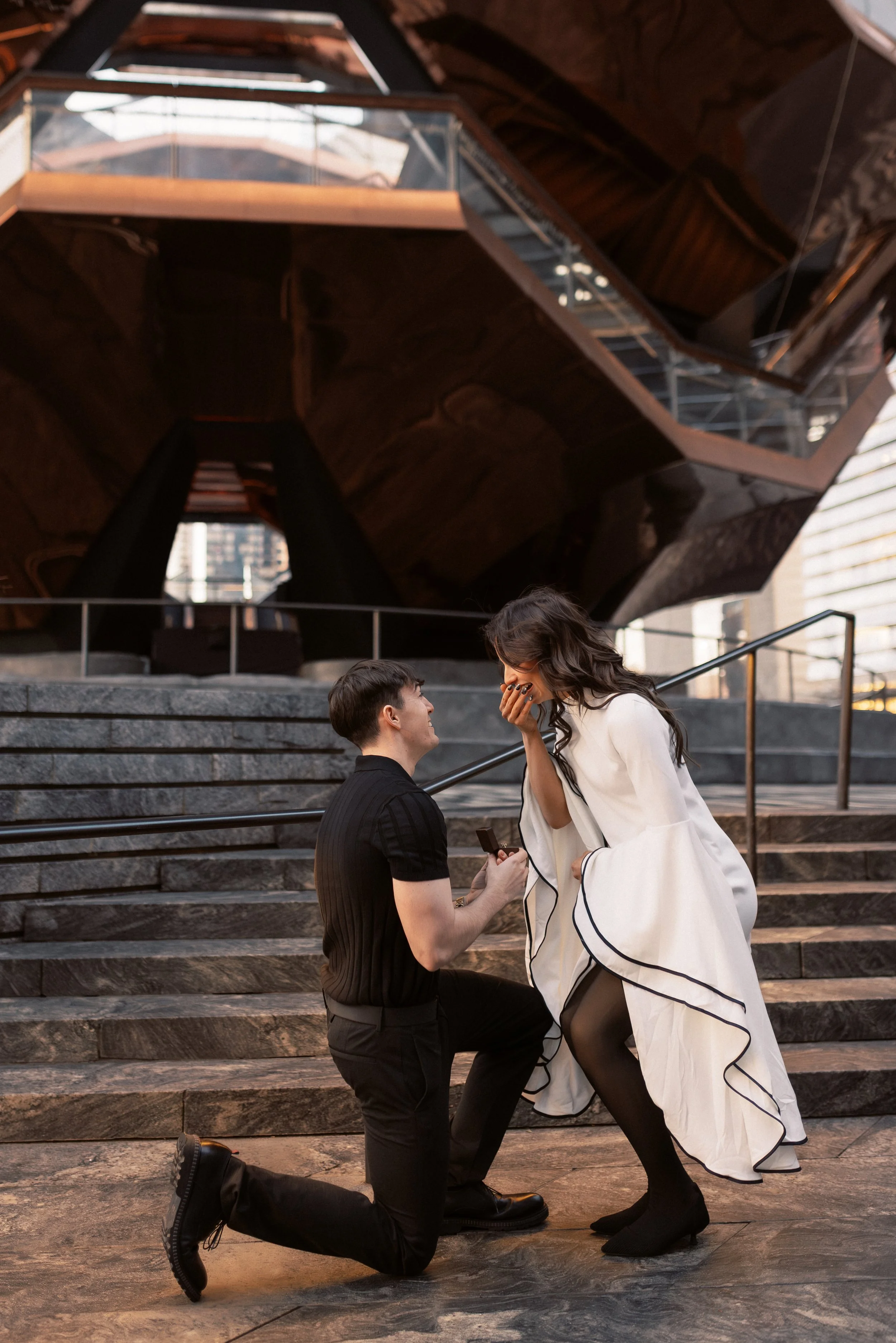 A man on one knee presenting a ring to a woman who appears surprised and is covering her mouth with her hand, with an architectural structure and stairs in the background.