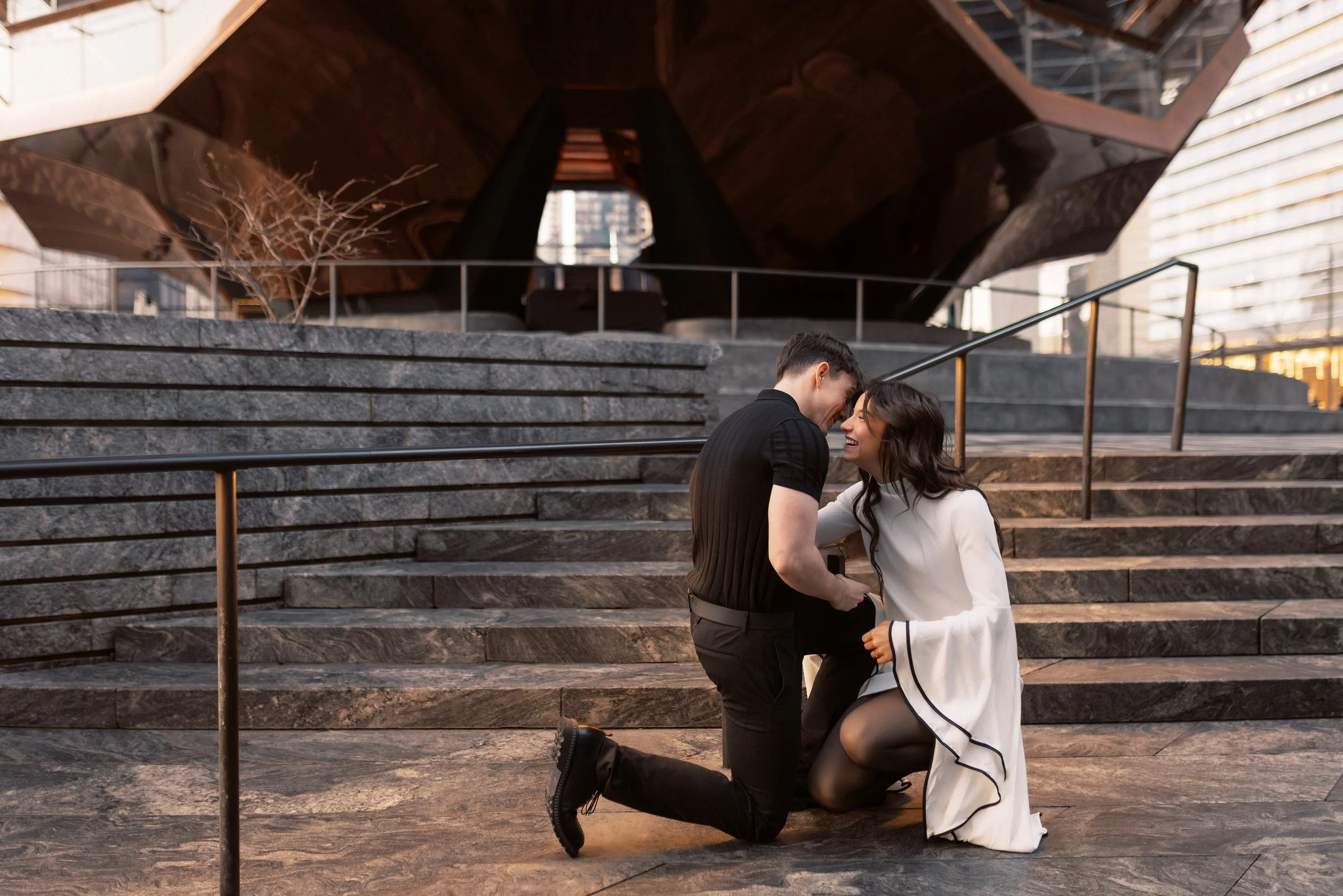 A man kneeling in front of a woman, holding her hand and both smiling, on a city staircase with modern buildings in the background.