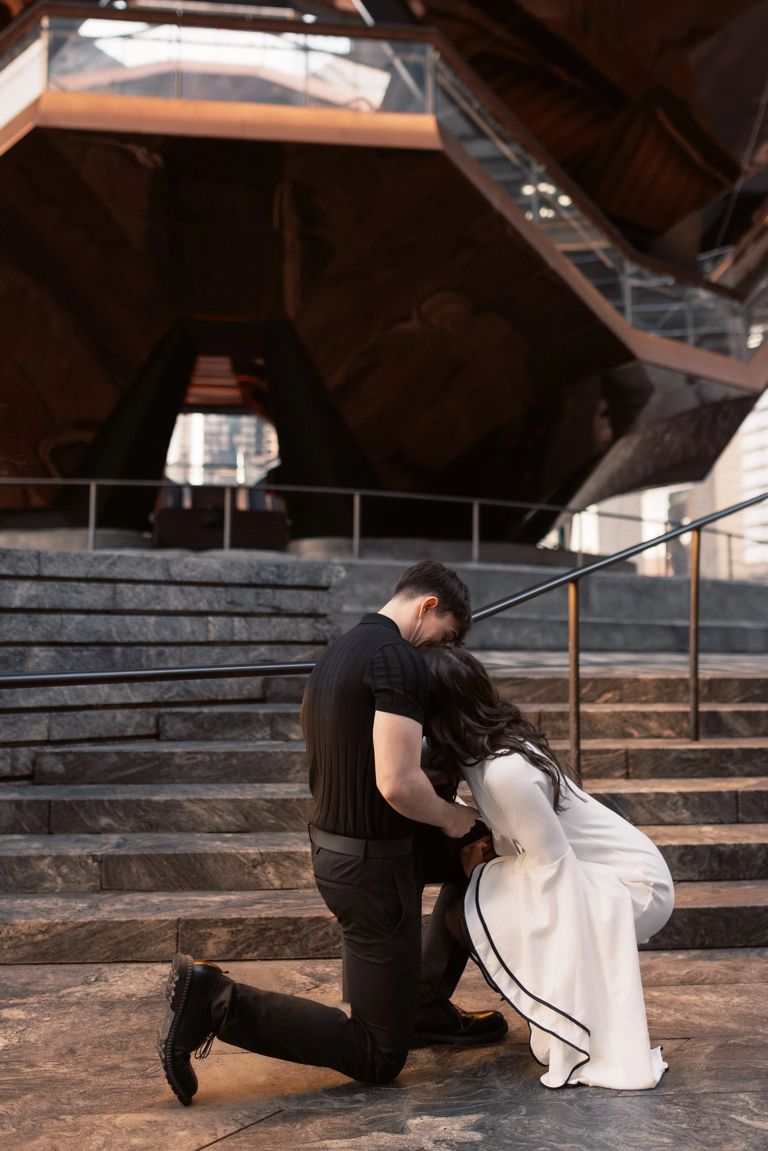 A man kneeling on one knee proposing marriage to a woman on a set of outdoor stairs. The woman is wearing a white dress and the man is dressed in a black shirt and pants. They are in front of a modern building with steps and metal railings.