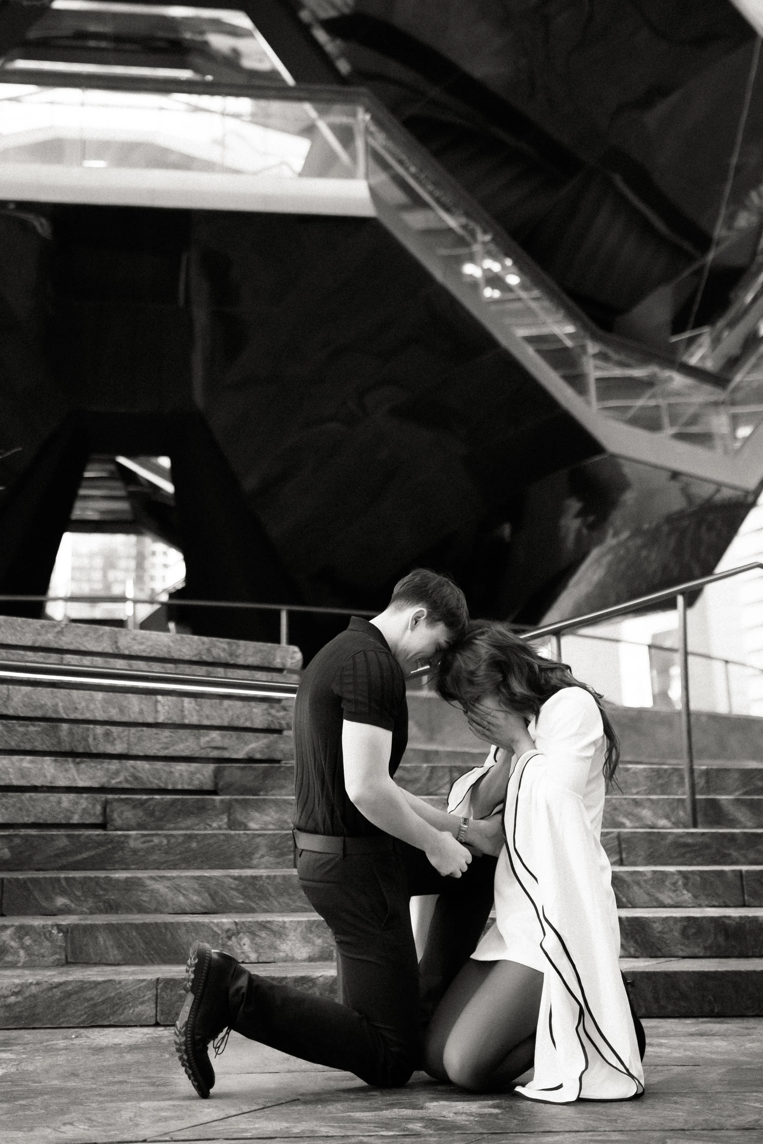 A man and a woman are kneeling on a staircase, holding hands, with the woman covering her face and the man looking at her. The setting is indoors with stairs behind them and a large, angular, reflective structure above.