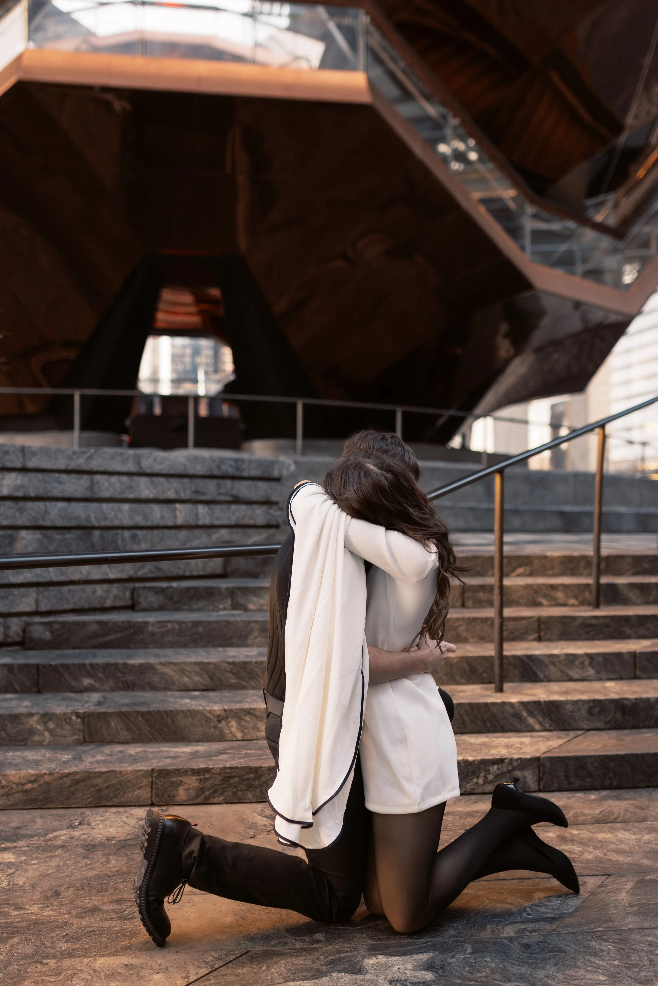 A couple dressed in casual and formal clothes is hugging and kneeling on a stone staircase outside a modern building with large windows and dark exterior. The woman has long hair and is wearing a white coat and tights, while the man has shorter hair 
