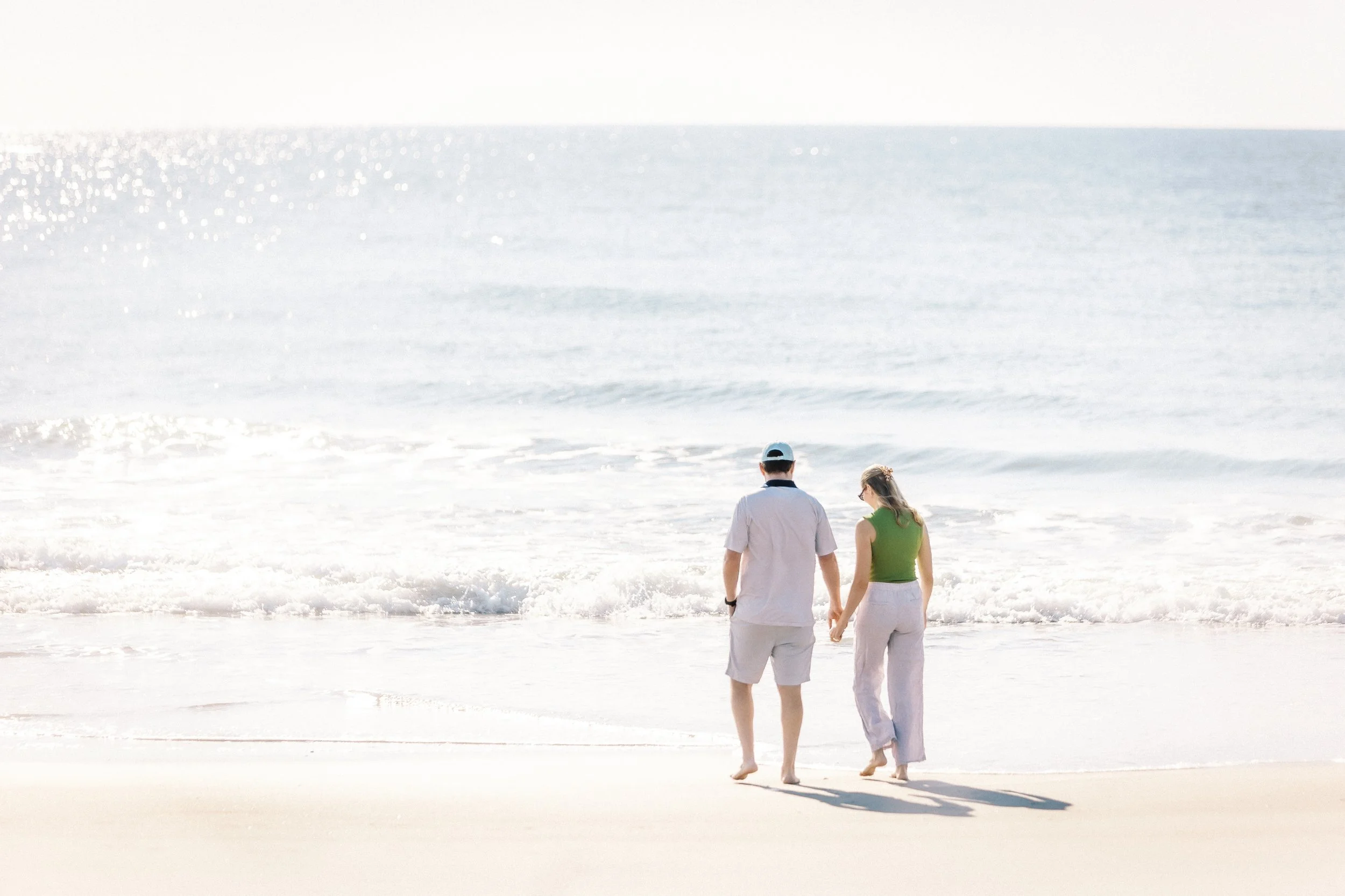 A couple walks along the beach holding hands with the ocean waves crashing at their feet under a bright, clear sky.