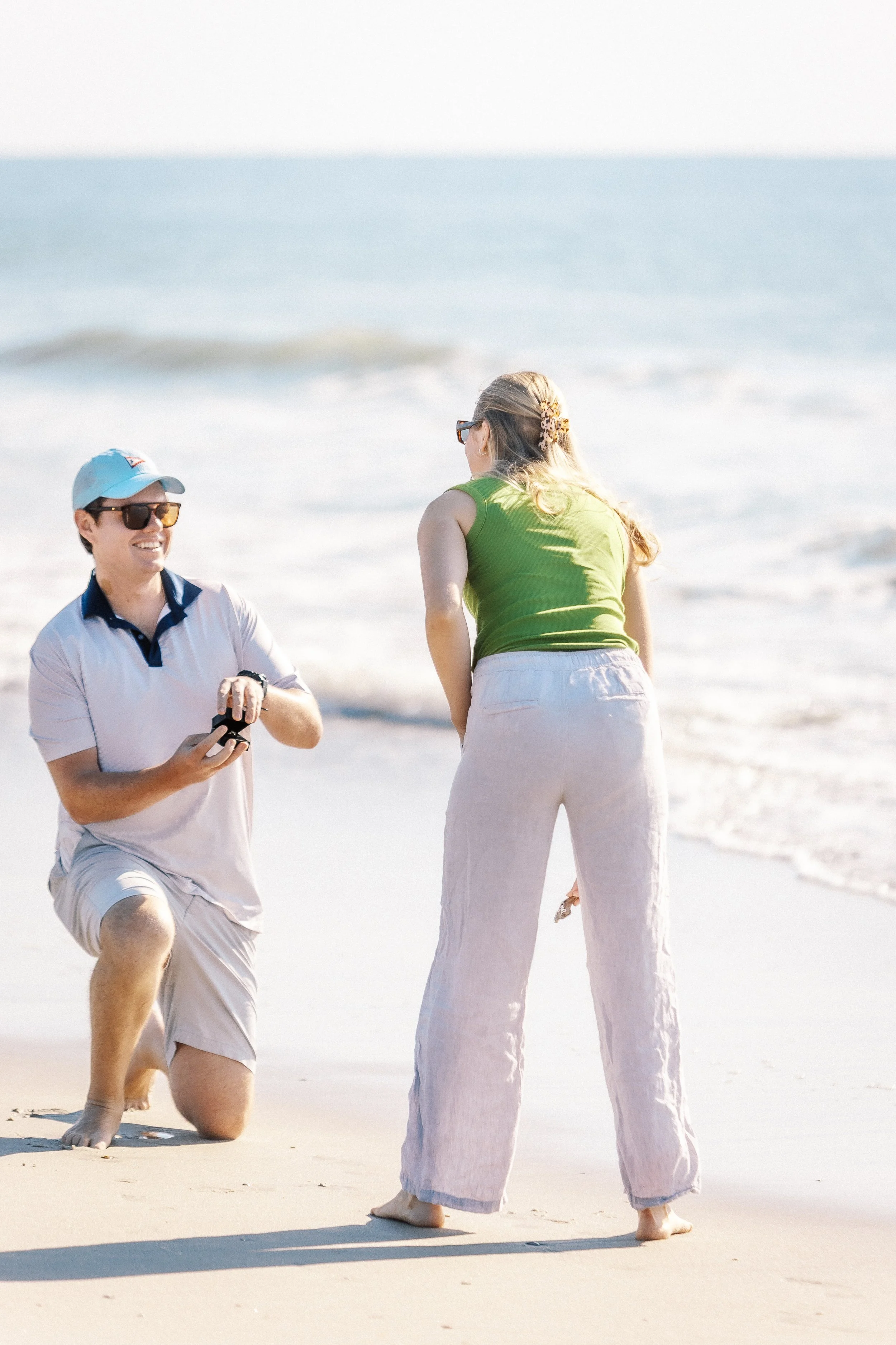 A man kneeling on one knee and a woman standing on a sandy beach with ocean waves in the background. The man is wearing sunglasses, a light blue cap, and a white polo shirt. The woman has long hair with a hair clip, glasses, a green sleeveless top, a