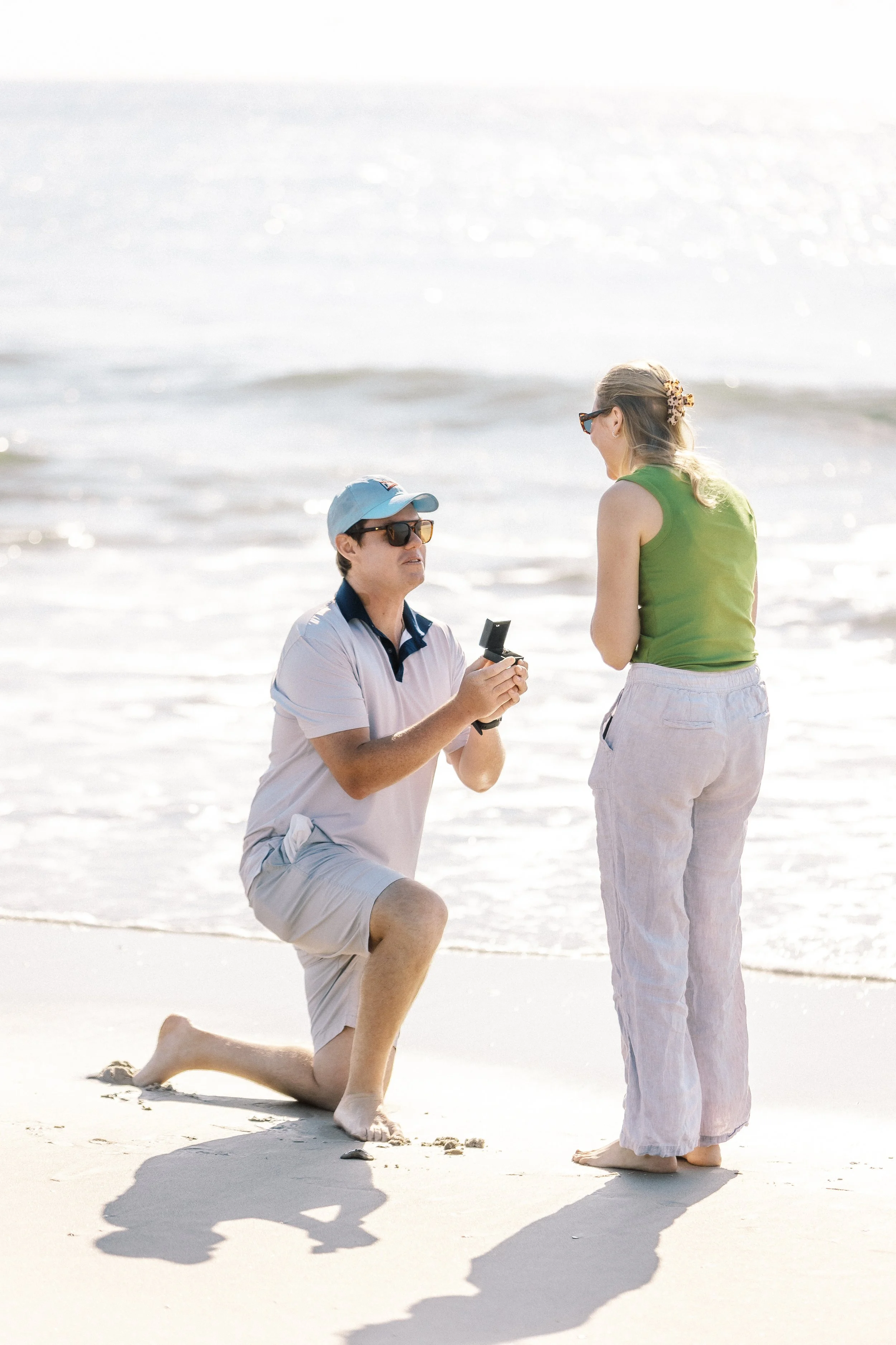 A man kneeling on one knee on the beach, holding a small box, proposing to a woman standing nearby in front of the ocean, both smiling.