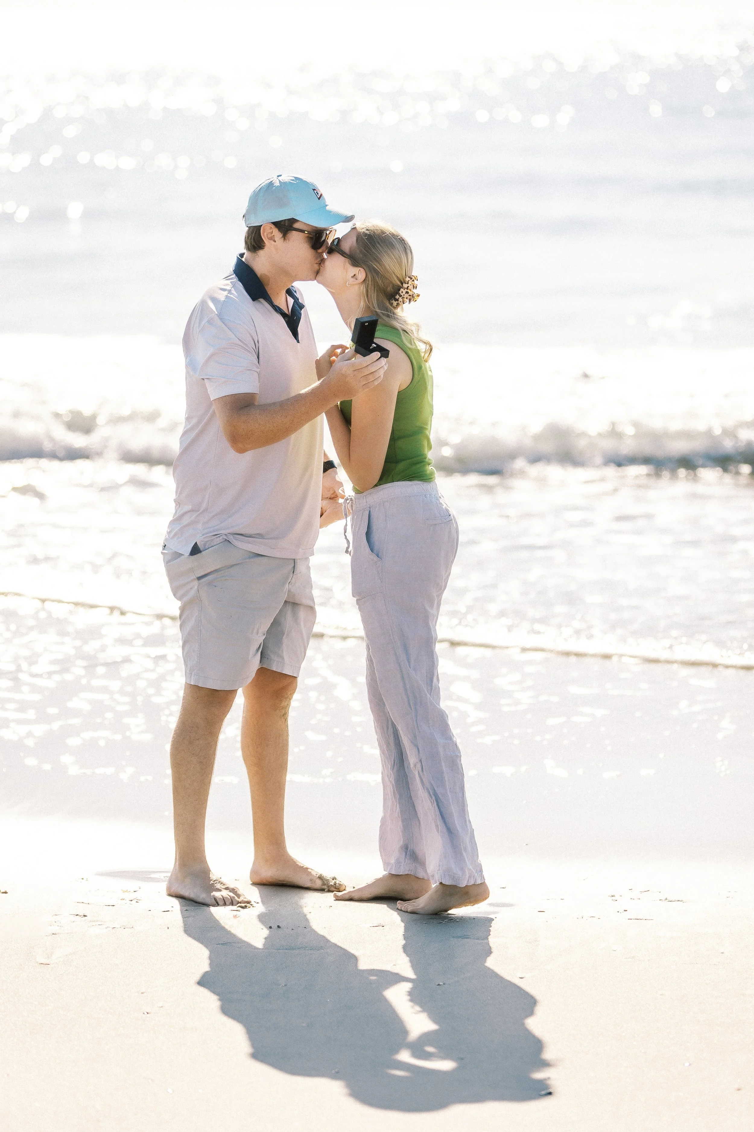 A couple stands on the beach, sharing a kiss with the ocean in the background. They are holding hands and looking at each other, with shadows cast on the sand.