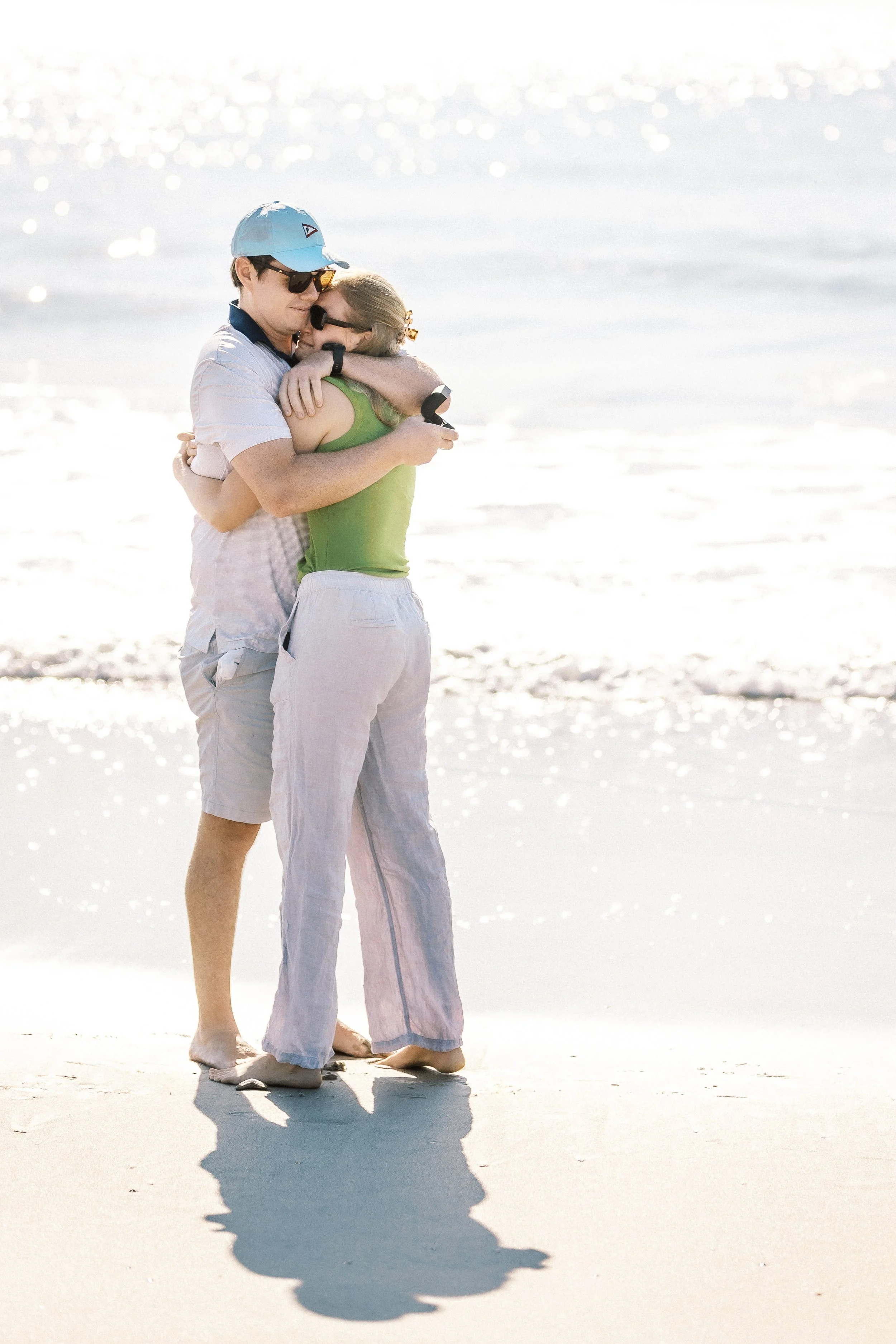 A couple hugging on the beach with bright sunlight reflecting off the water in the background.