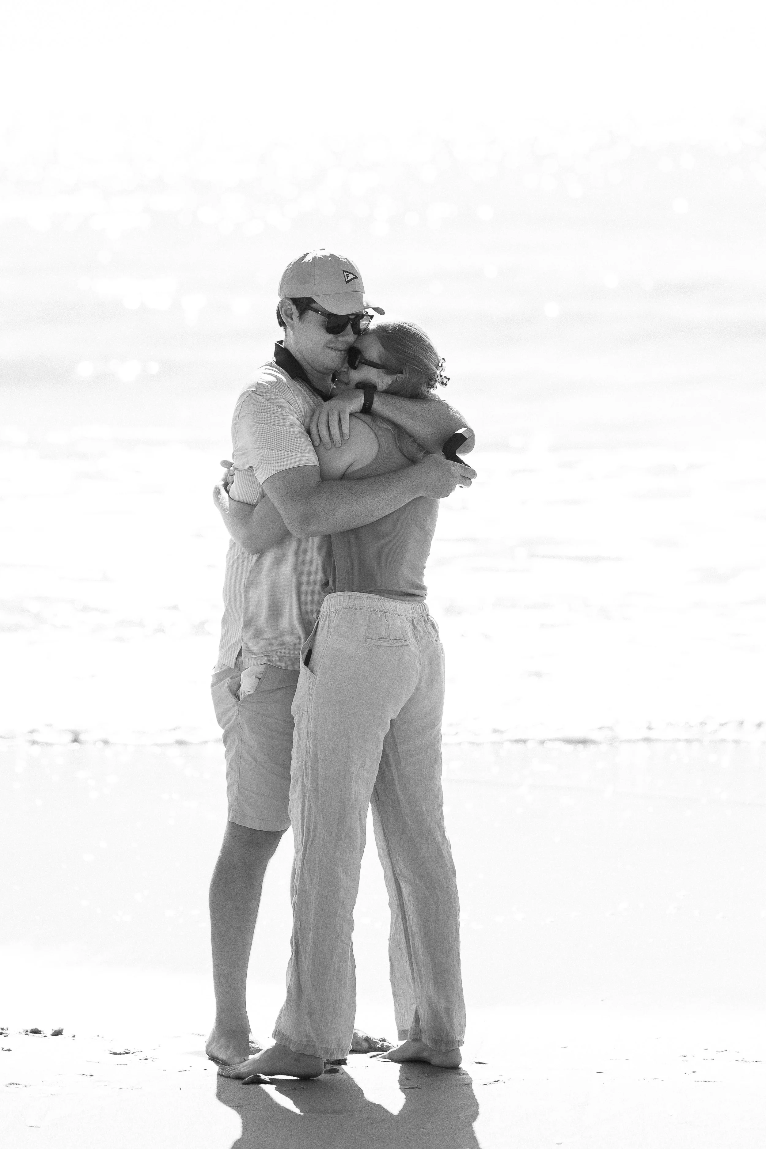 A couple hugging on a beach with the ocean in the background and the sun creating a bright reflection on the water.