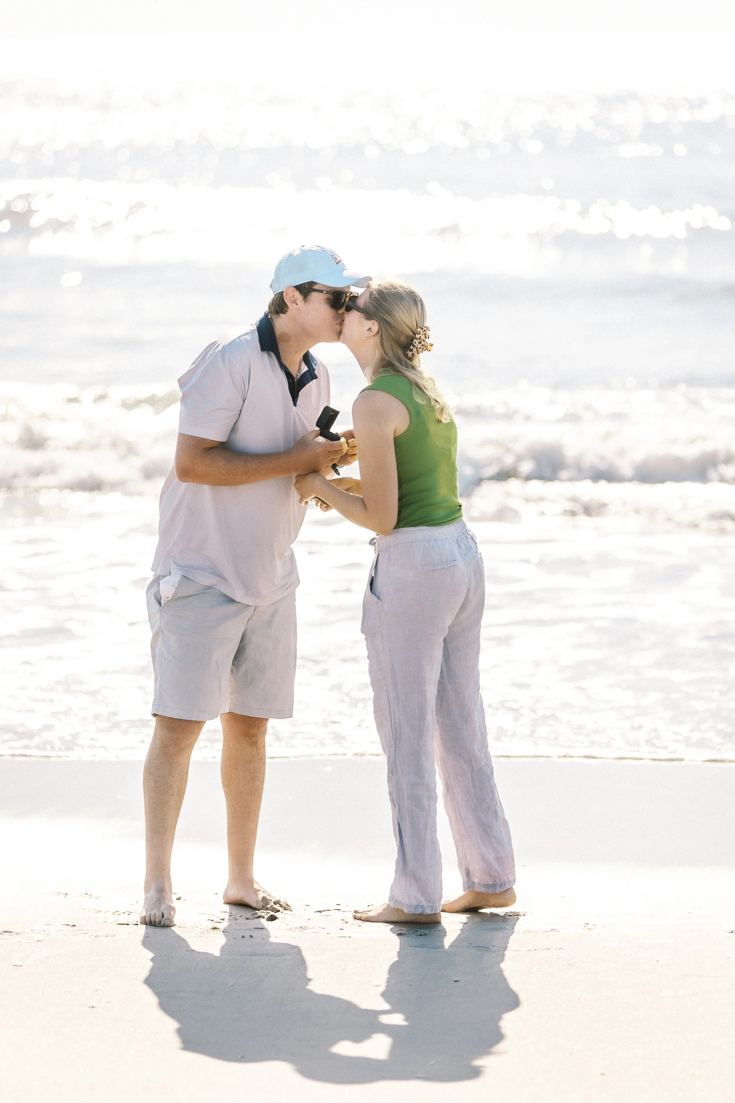 A couple kissing on the beach with the ocean in the background, both wearing casual summer clothes and sunglasses.