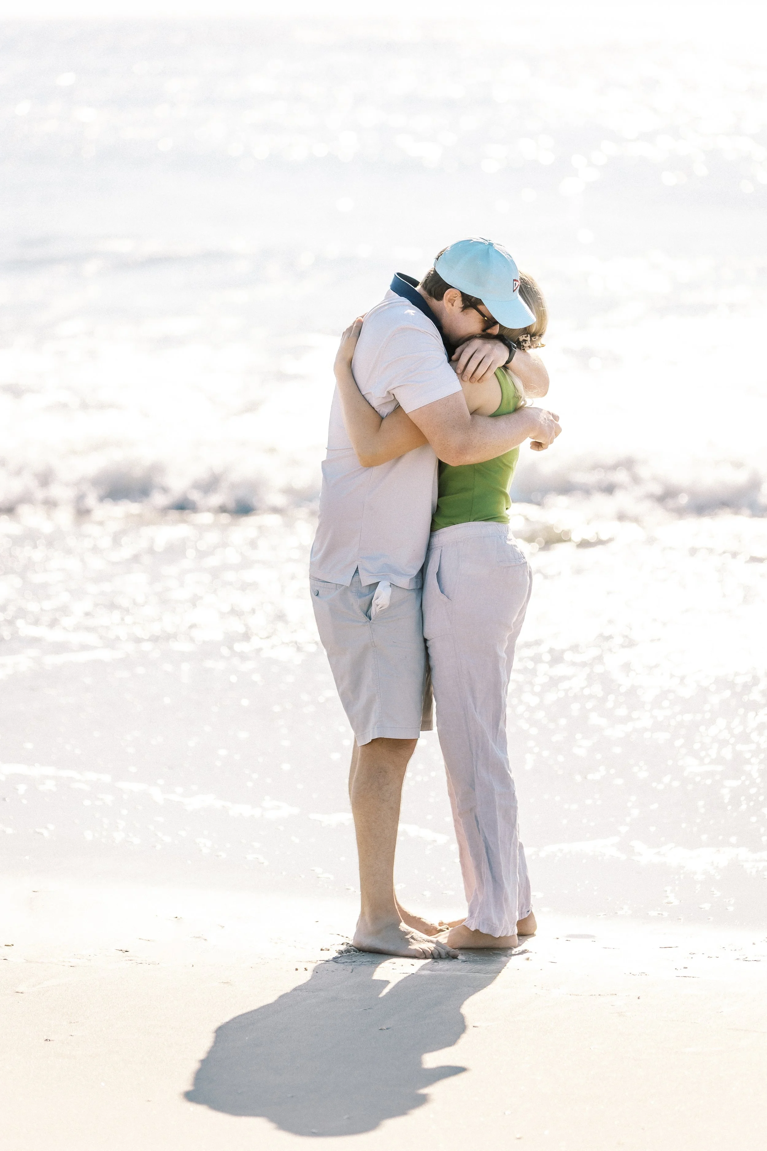 A couple hugging on a sandy beach with gentle waves, both barefoot, with sunlight reflecting off the water.