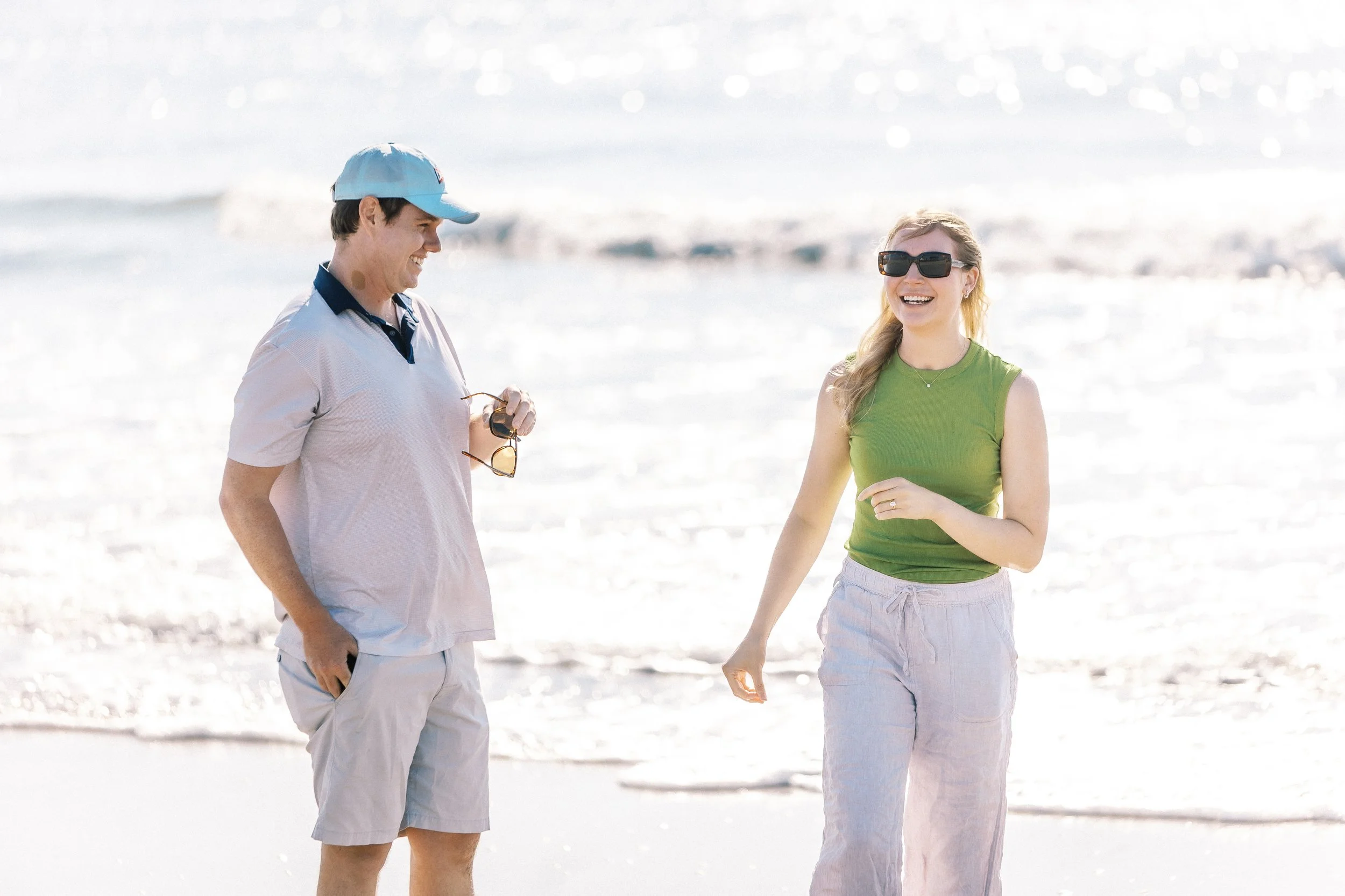 Two people, a man and a woman, enjoying a conversation on a sunny beach. The man wears a light polo shirt, shorts, and a cap, while the woman wears sunglasses and a green top with light pants. They are smiling and appear happy.