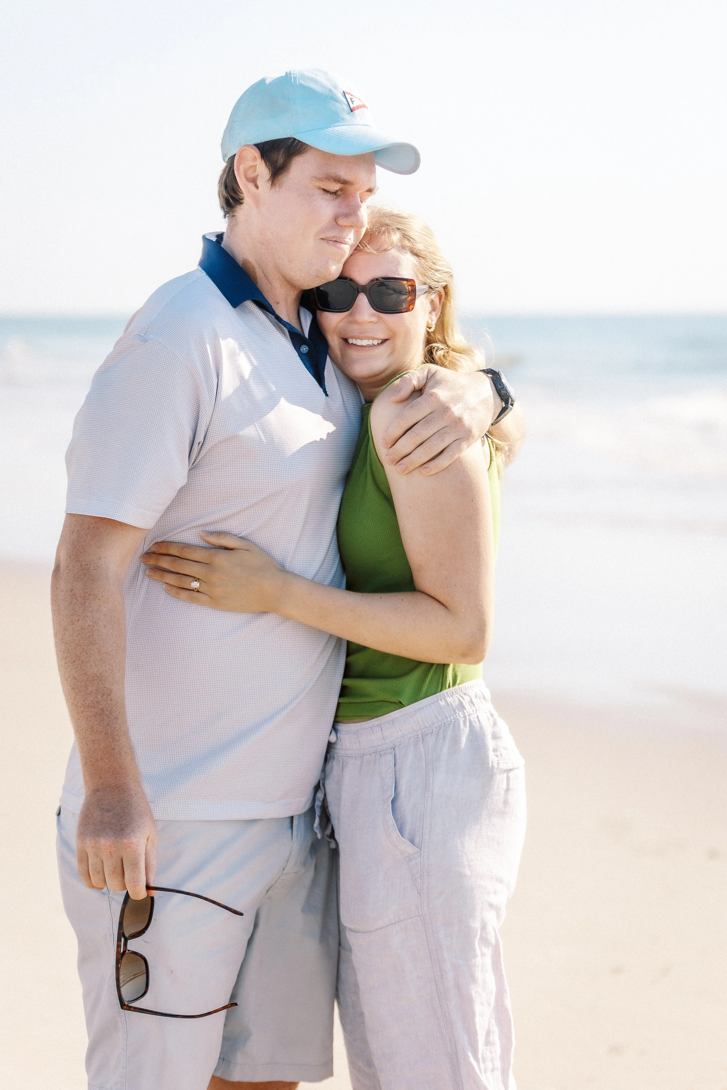 A couple hugging on a beach, smiling, with the ocean in the background.