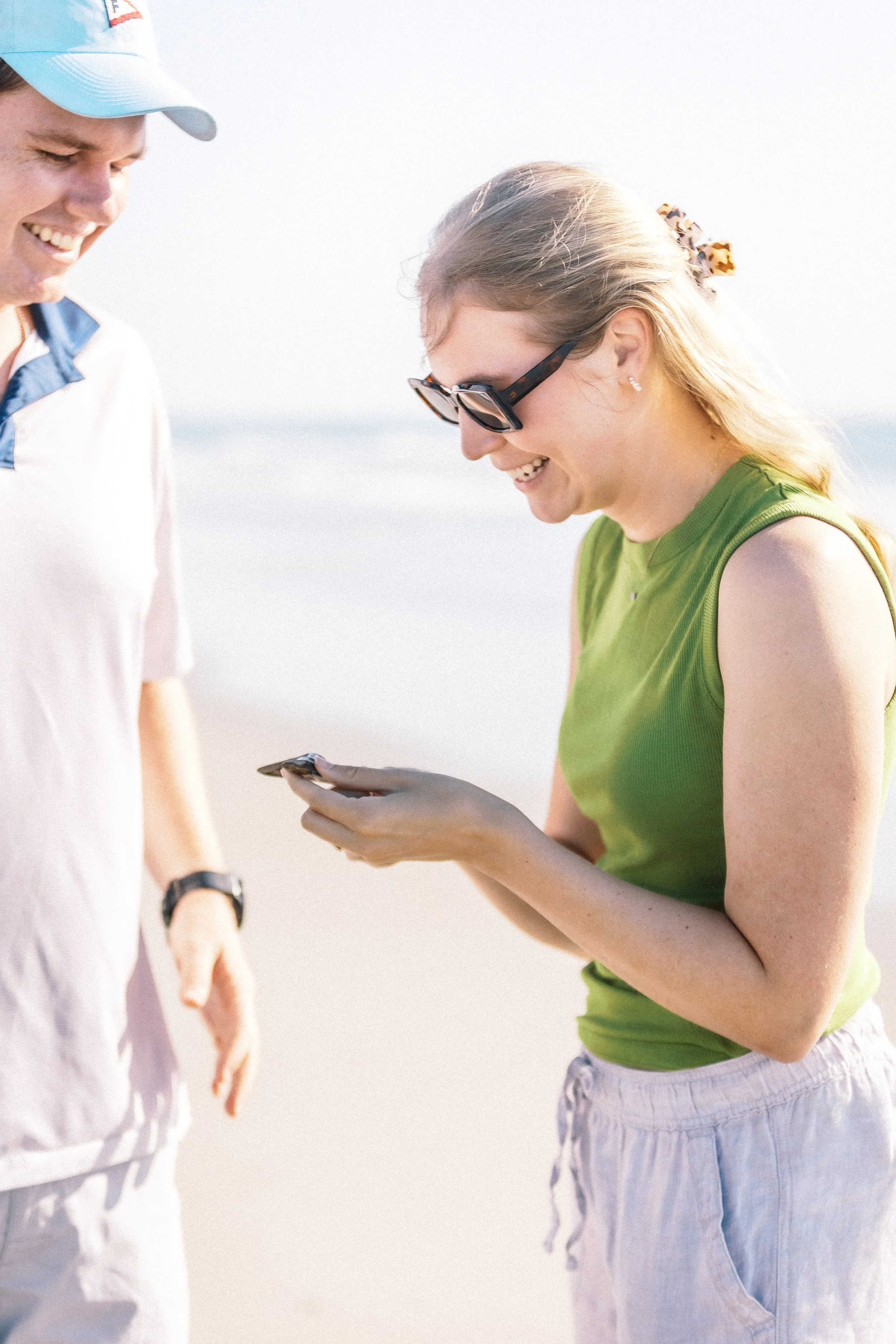 A woman in a green sleeveless top and sunglasses showing something on her phone to a smiling man on a beach.