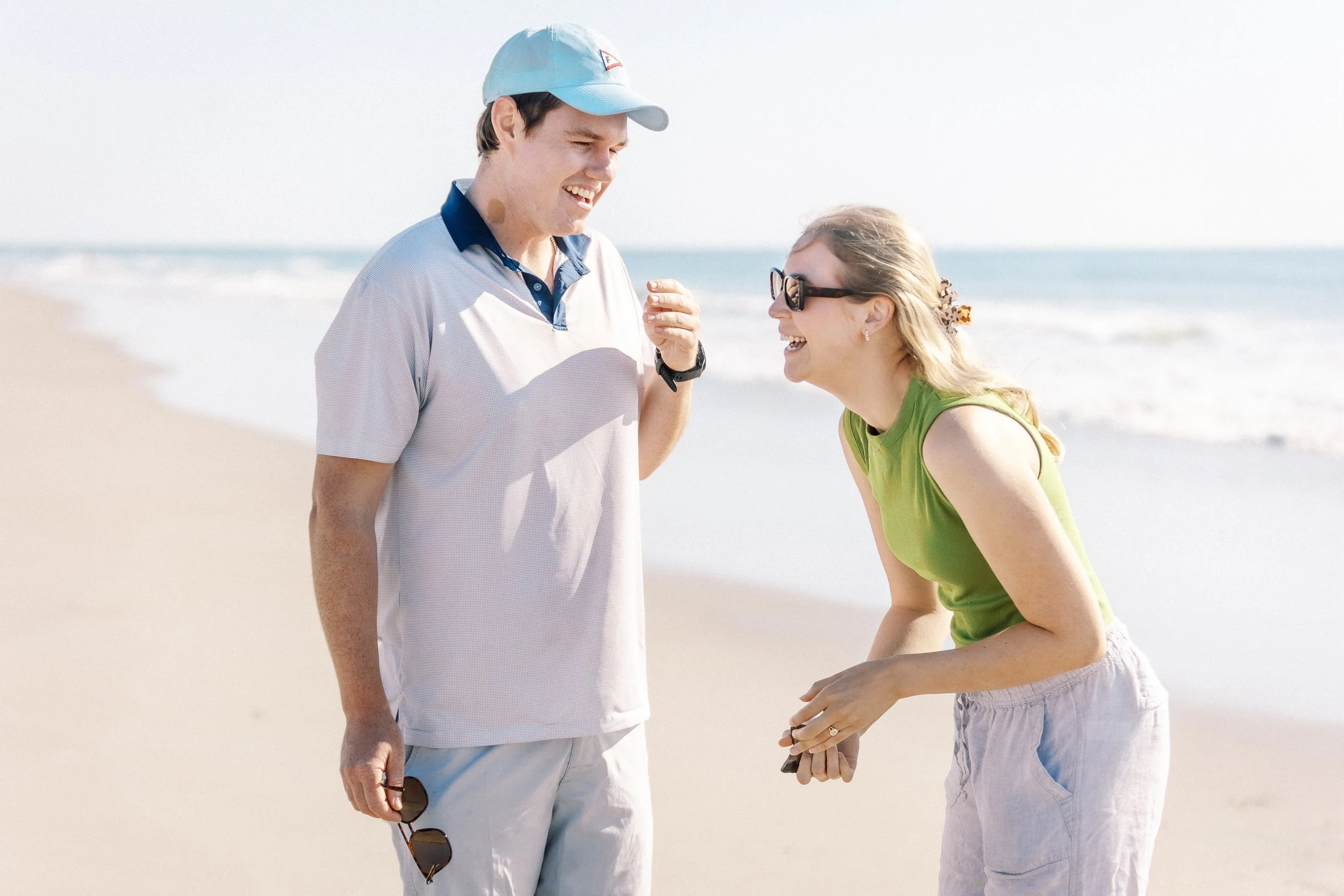 A man and a woman are standing on a beach, smiling and laughing while talking to each other. The man wears a light blue cap and a short-sleeved shirt, holding sunglasses in his right hand. The woman wears sunglasses, a green sleeveless top, and light