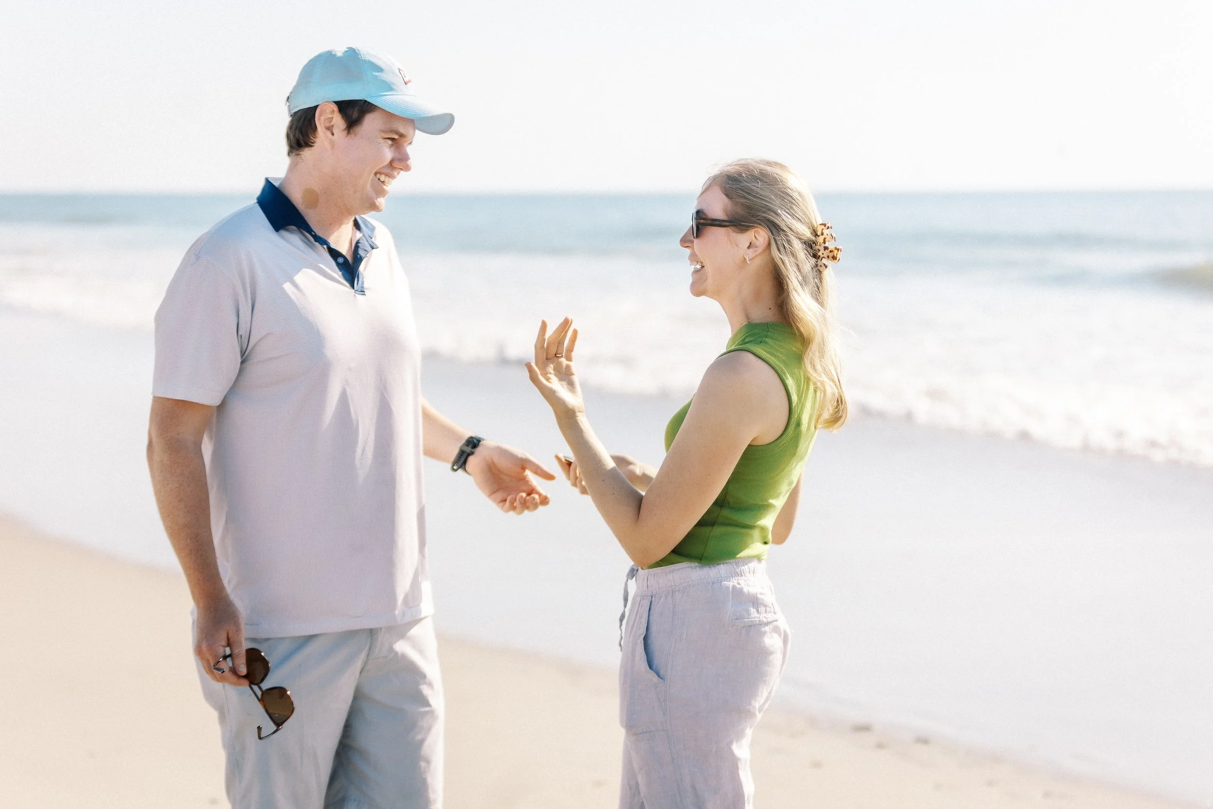 Two people, a man and a woman, are smiling and talking at the beach with the ocean in the background. The man is wearing a light-colored polo shirt, a light blue cap, and holding sunglasses, while the woman is wearing sunglasses, a green tank top, an