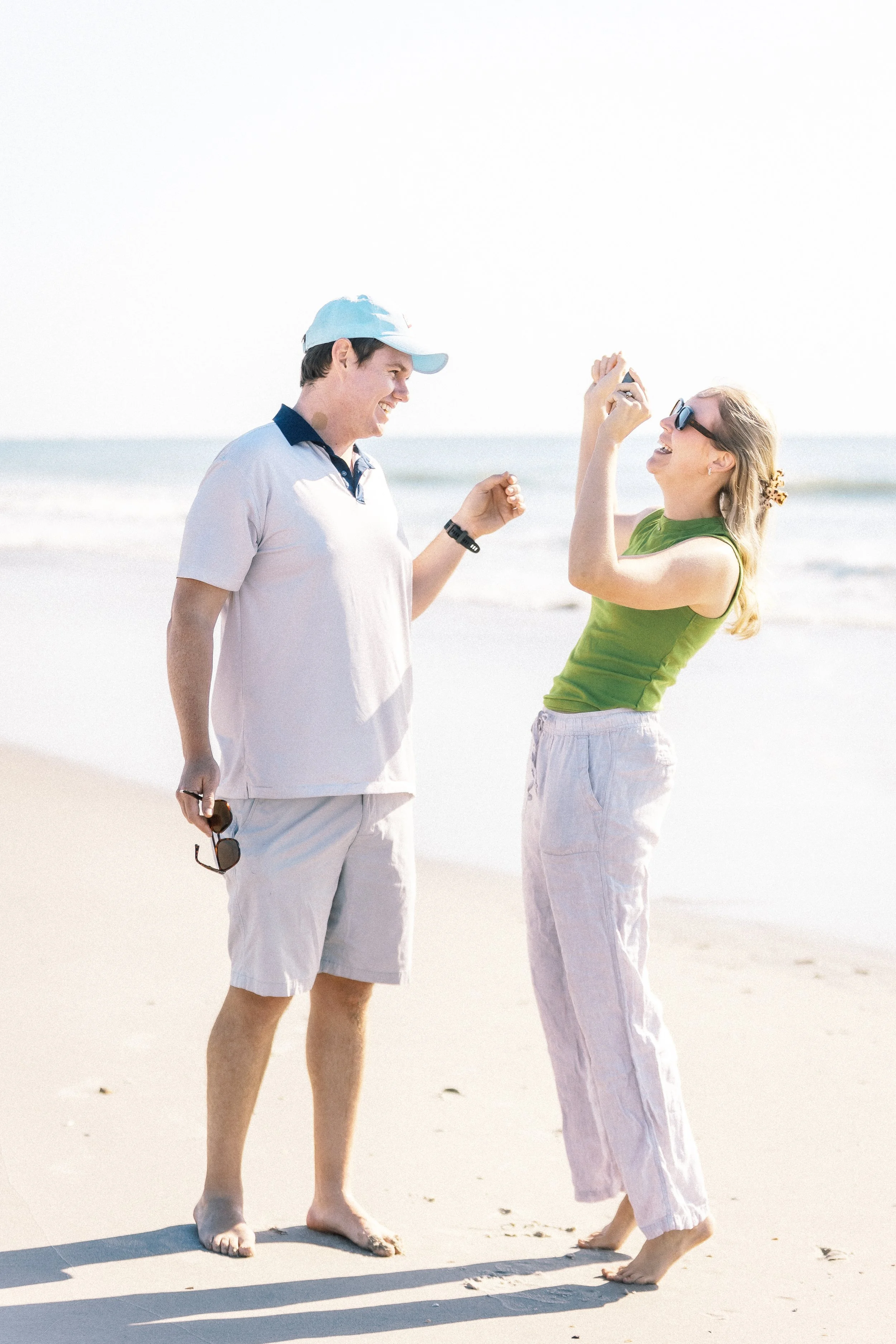 A man and woman are at the beach, smiling and laughing. The man wears a light-colored polo shirt, shorts, a light blue cap, and is holding sunglasses. The woman wears sunglasses, a green sleeveless top, and light-colored pants, with a hair tie with a