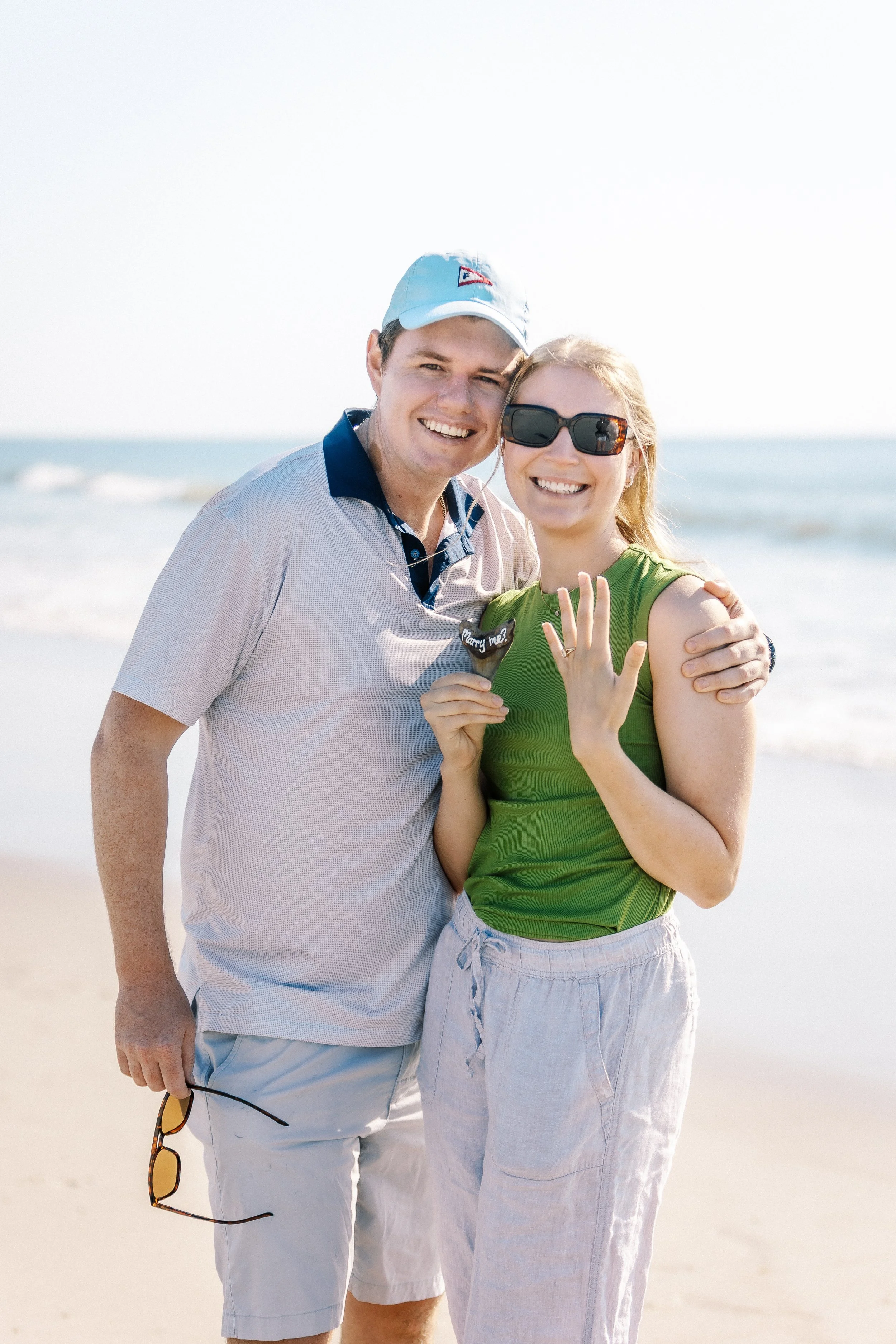 A happy couple standing on the beach, smiling, with the ocean in the background. The woman is showing off an engagement ring.