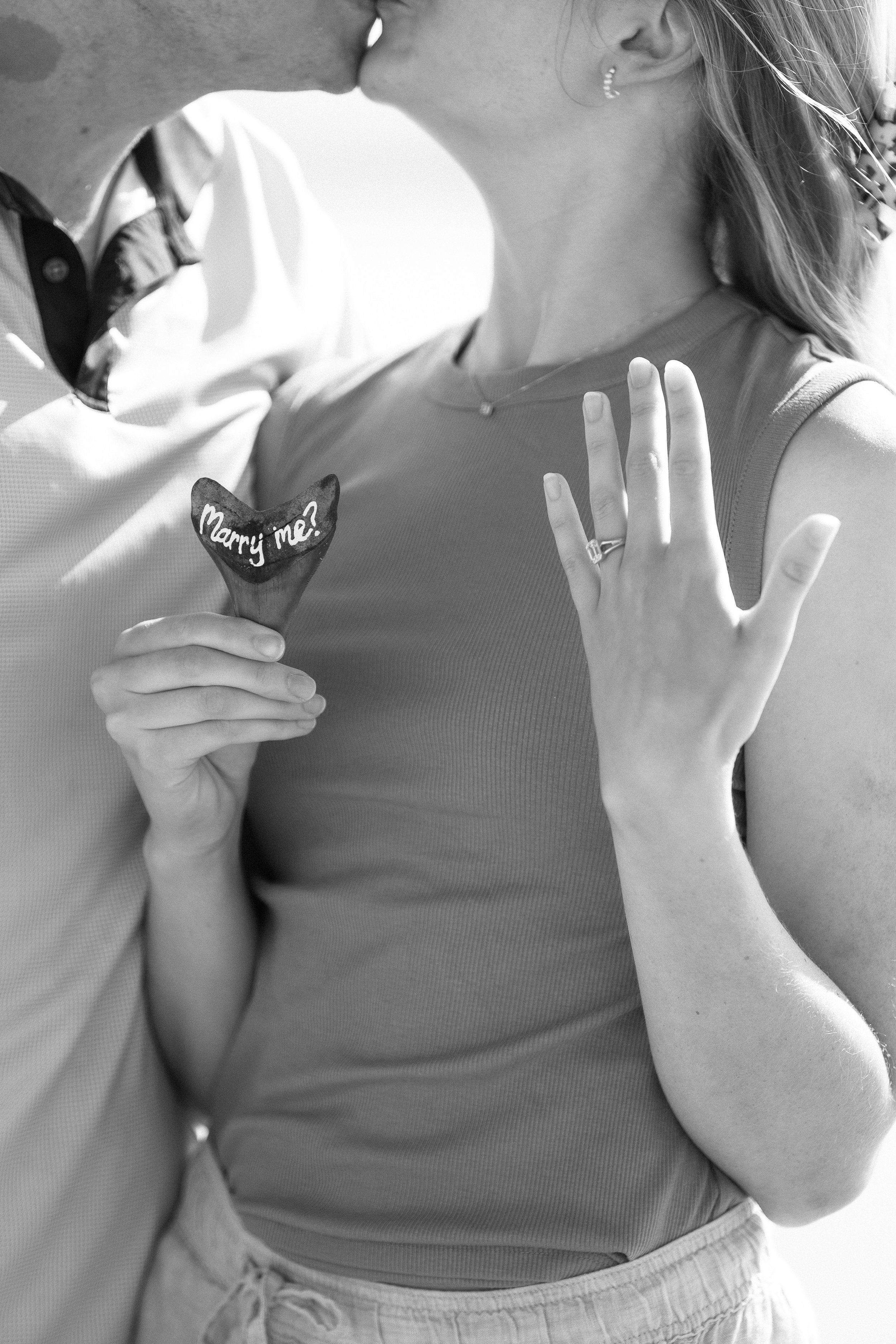 A couple sharing a kiss, with the woman holding a heart-shaped chocolate with 'Marry me?' written on it, and the woman's left hand displaying a ring.