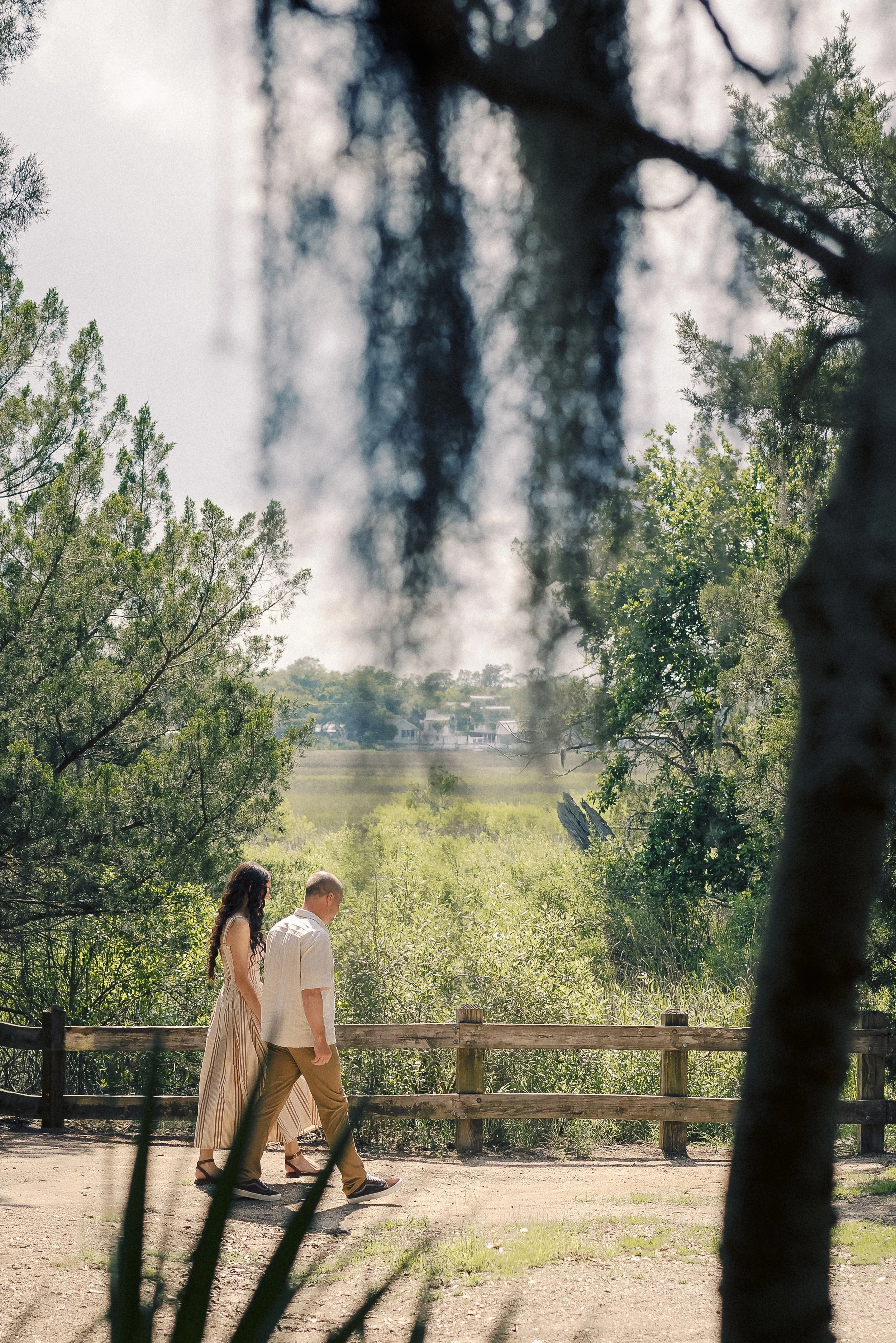 A couple walking together on a path in a park, surrounded by trees and greenery, with a foggy background and some houses visible in the distance.