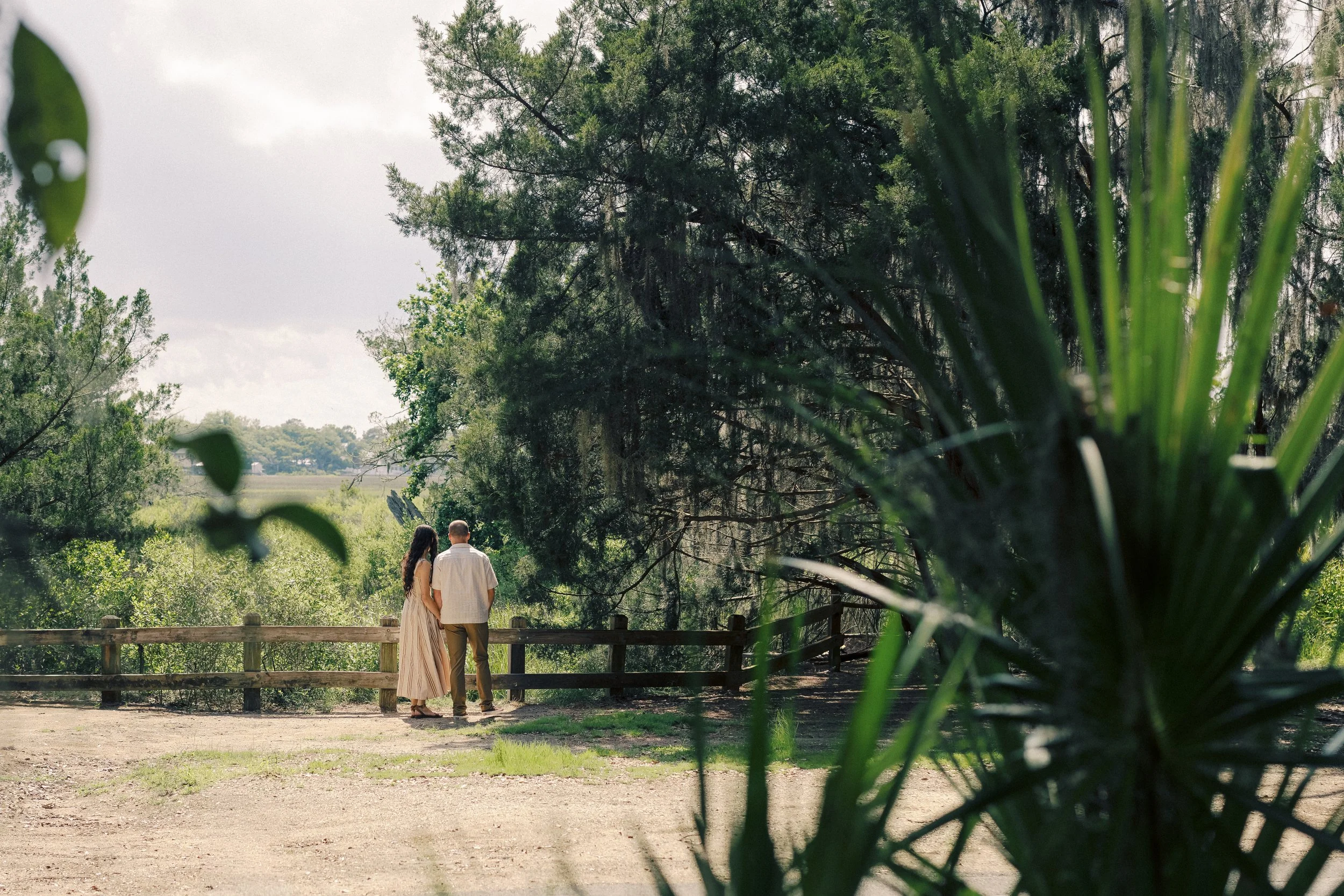 A couple walking together in a park surrounded by trees and greenery, with a wooden fence and open field in the background.