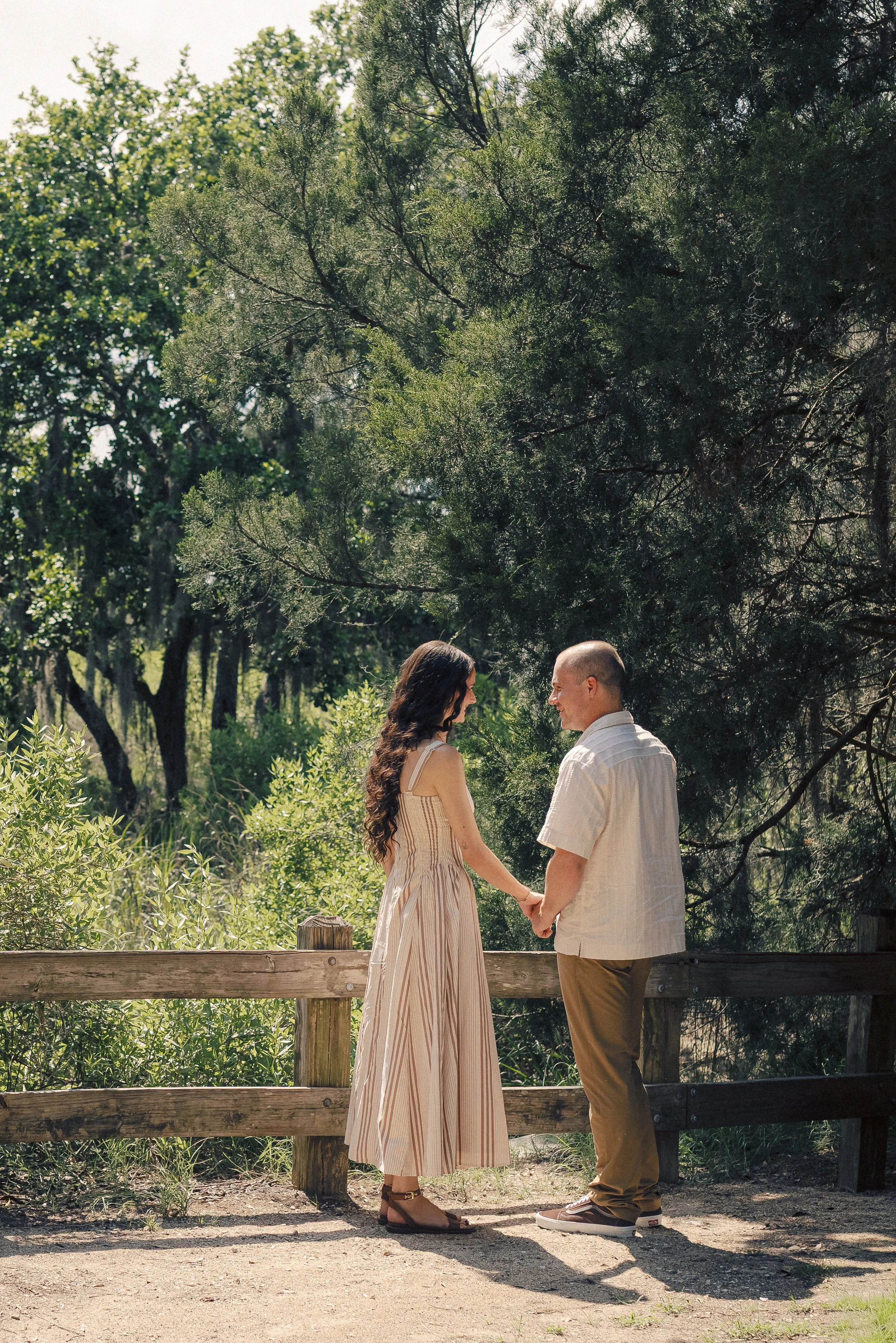 A couple holding hands and standing face to face on a wooden platform in a lush park with trees.